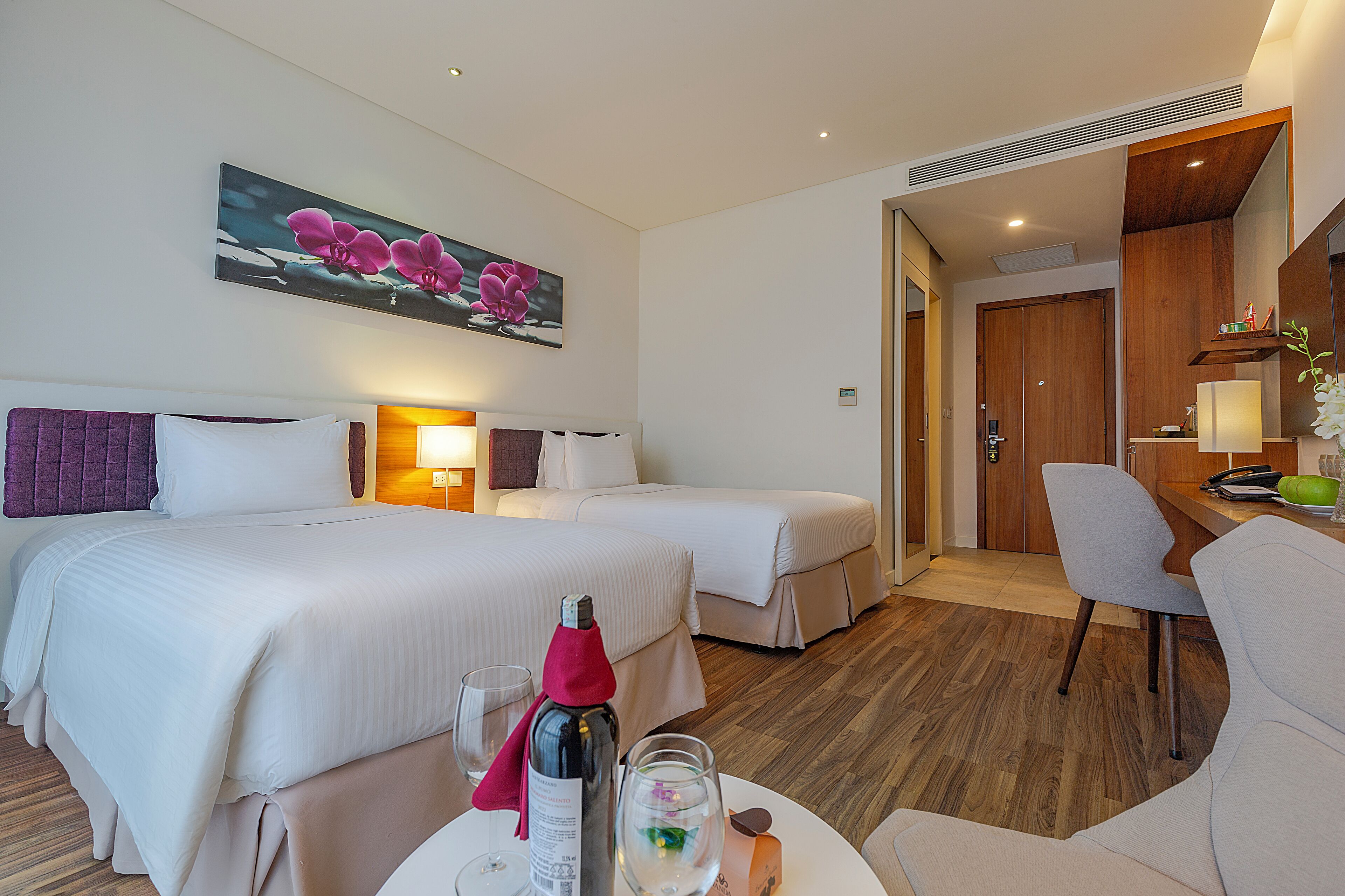 Quarto de hotel com duas camas, uma escrivaninha e uma cadeira. Há uma pintura na parede, uma garrafa de champanhe e copos na mesa.