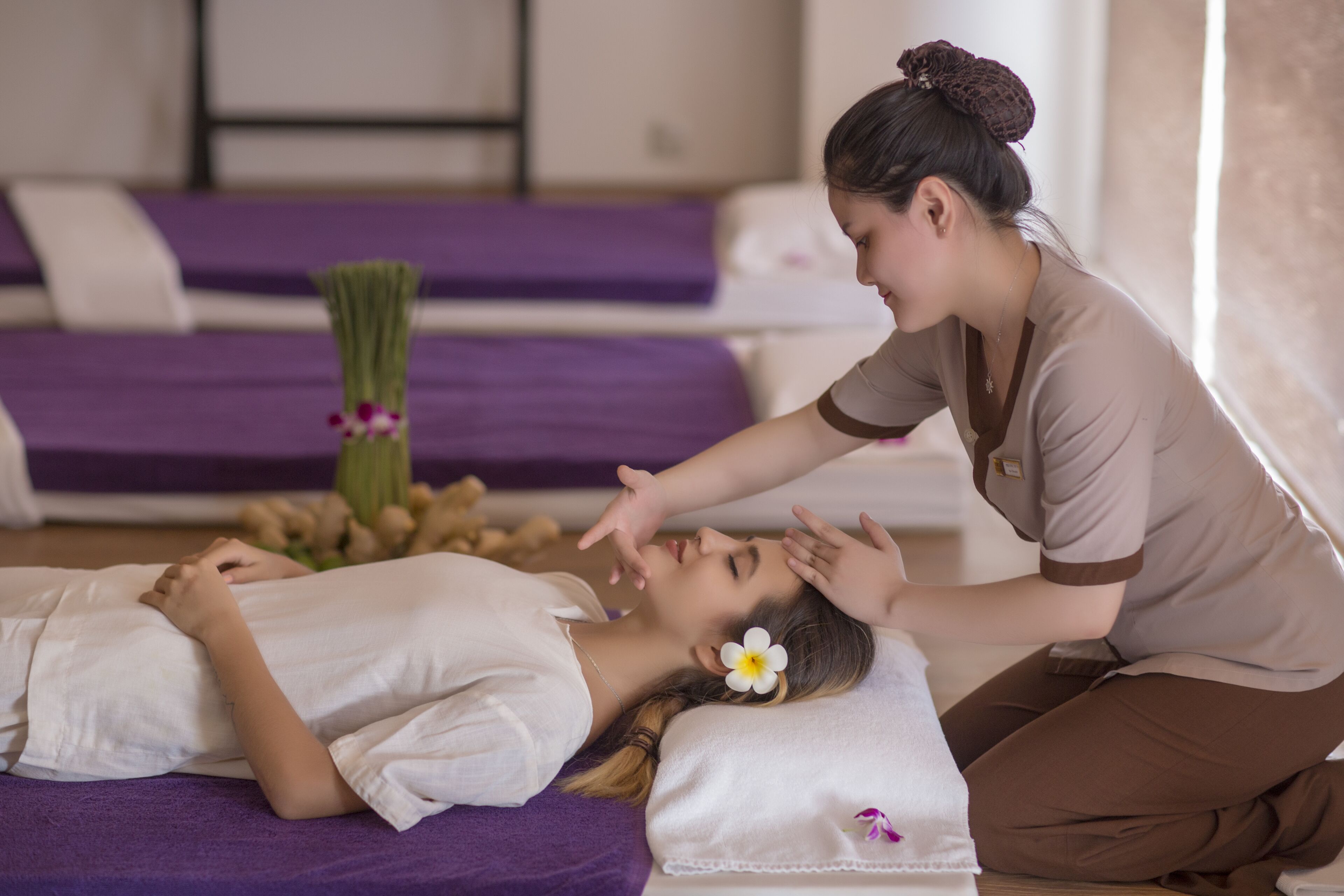 A person lying on a massage table in a room with purple bedding and a small plant.