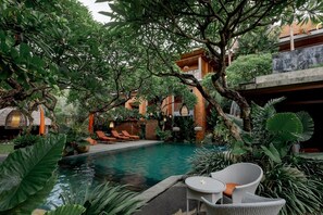 A pool area with lounge chairs, surrounded by lush greenery and a building with a thatched roof. - Fourteen Roses Boutique Hotel Legian (Kuta)