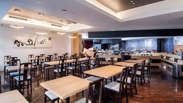 A spacious dining area with multiple tables and chairs, a kitchen in the background, and a modern ceiling design.