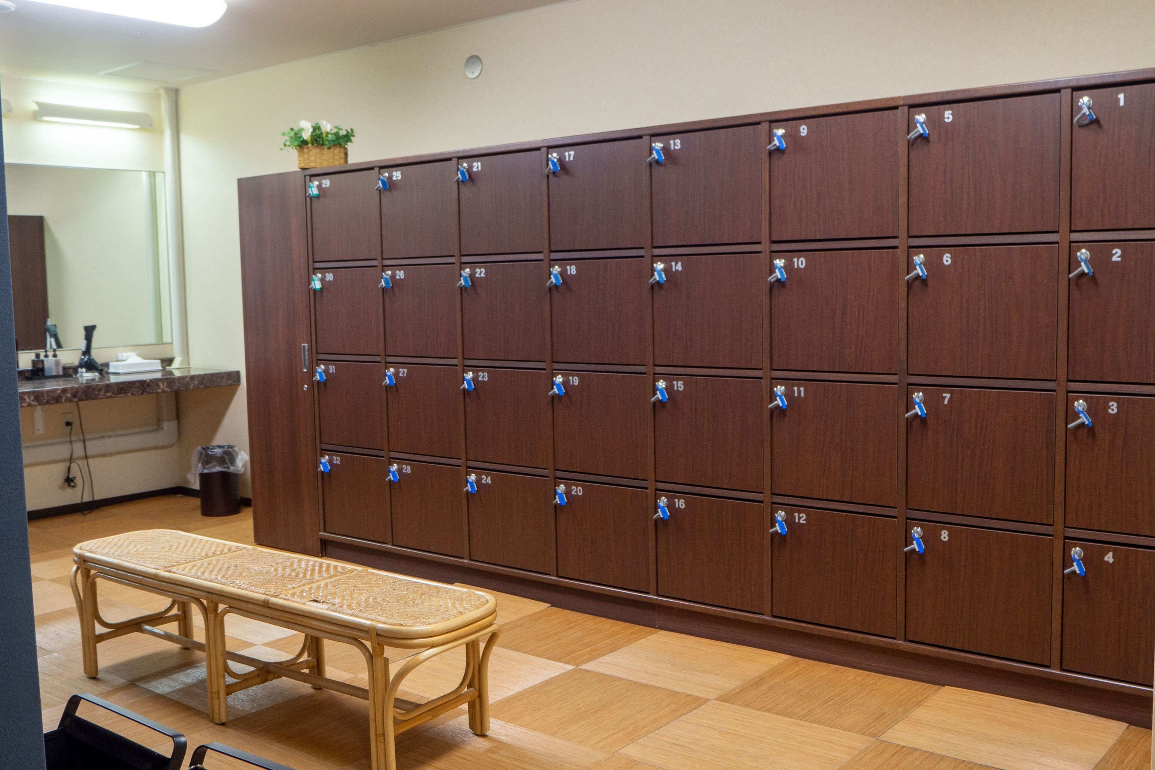 A row of wooden lockers with numbered locks, a bench, and a trash can in a room with a tiled floor.