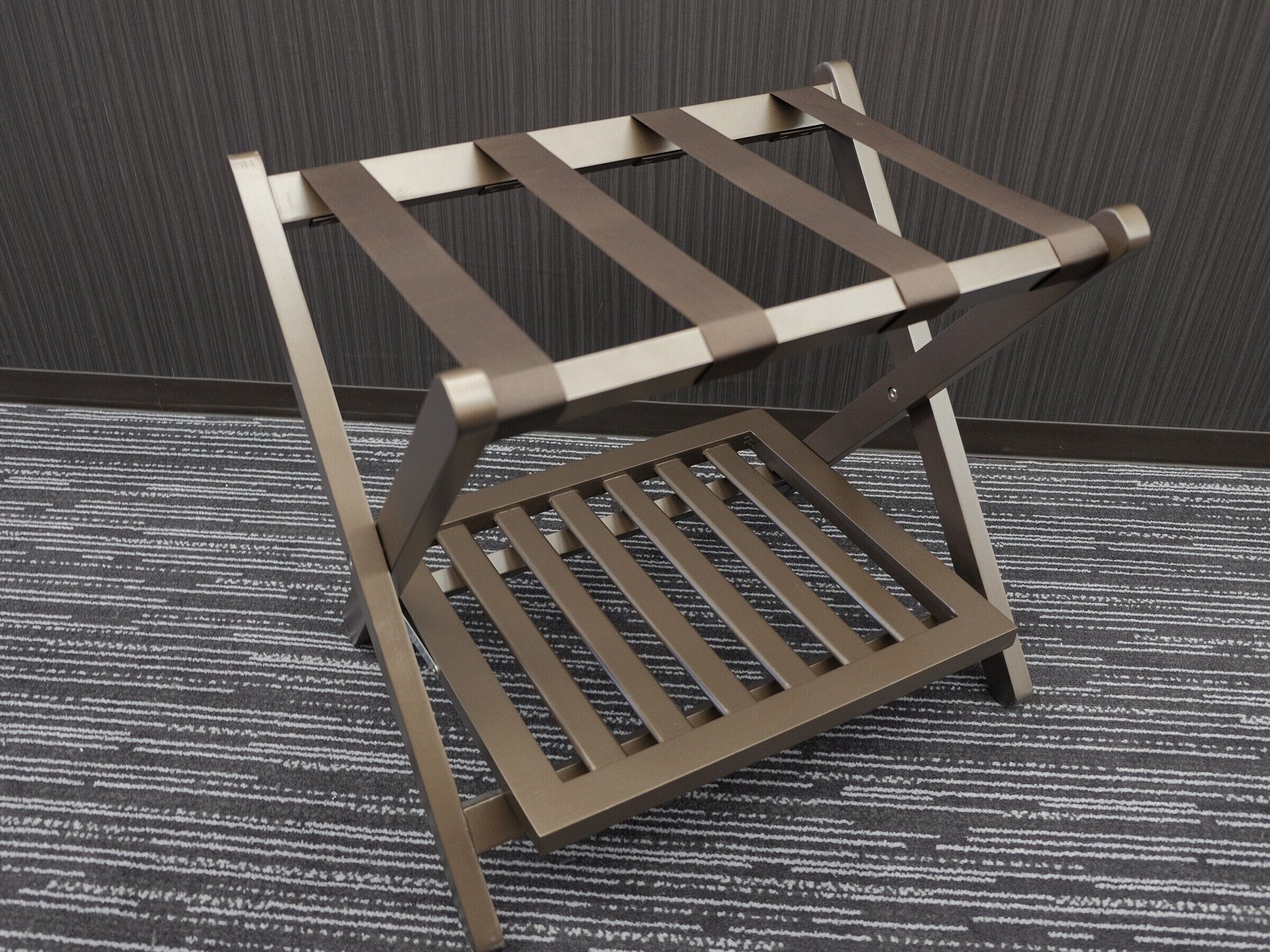 A metal chair with a slatted seat and backrest, positioned on a carpeted floor against a wall.
