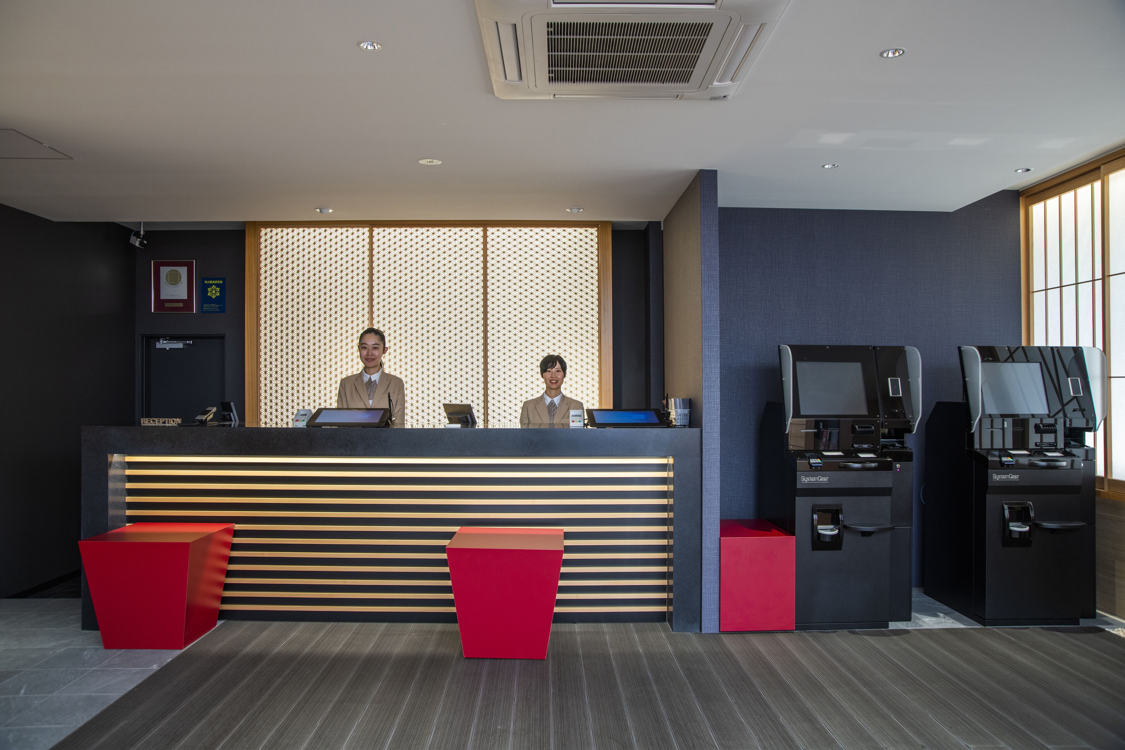 A reception area with a modern design, featuring a reception desk, two receptionists, and a seating area with red chairs.