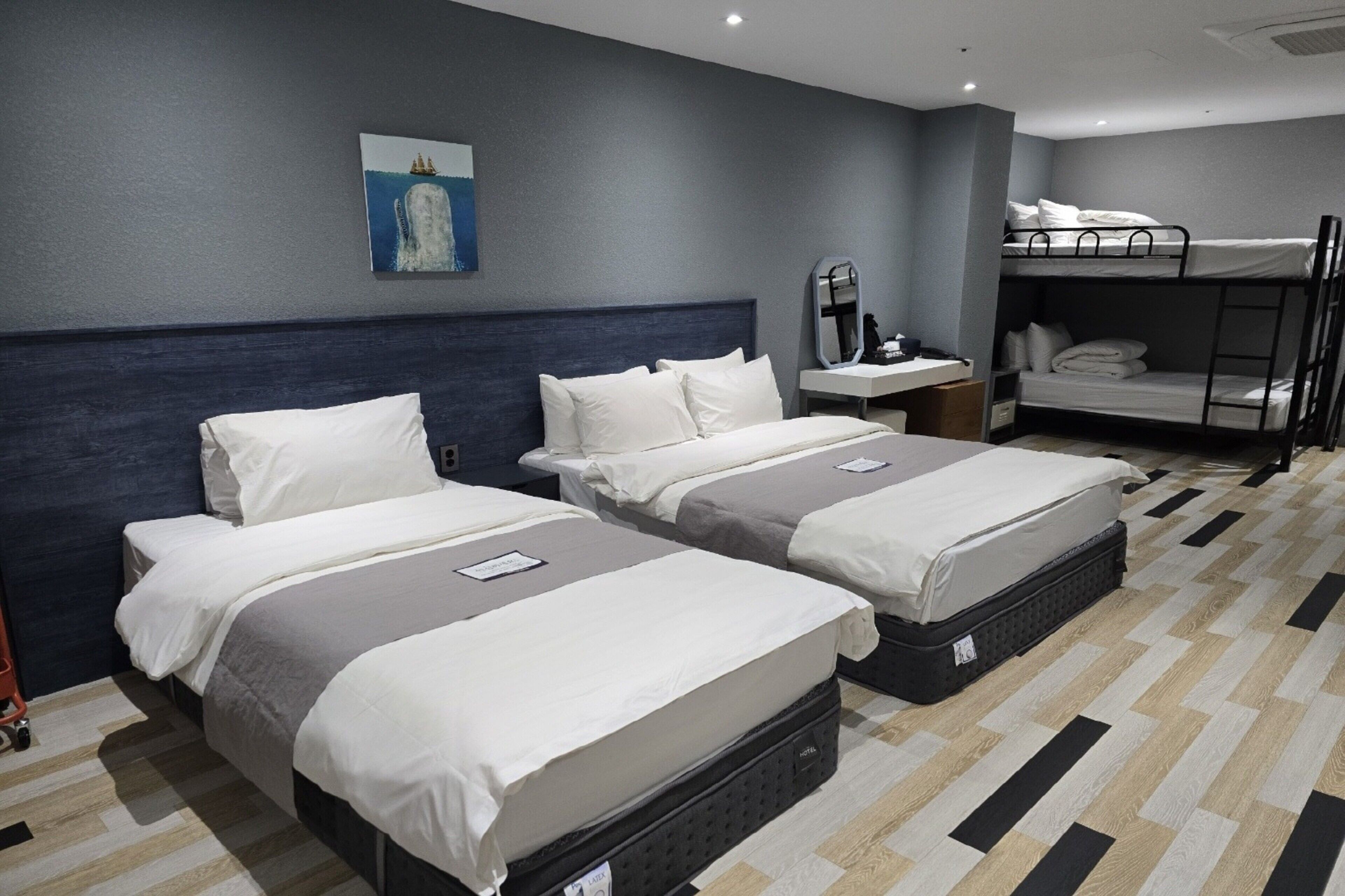 A hotel room with two beds, a bunk bed, a bedside table with a mirror, and a painting on the wall.