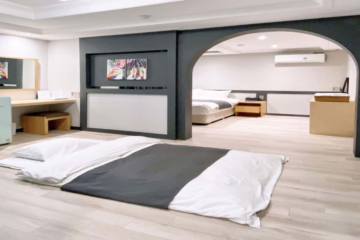 A modern hotel room with a large bed, a desk, and a wardrobe. There is a painting on the wall and an air conditioning unit above the bed.