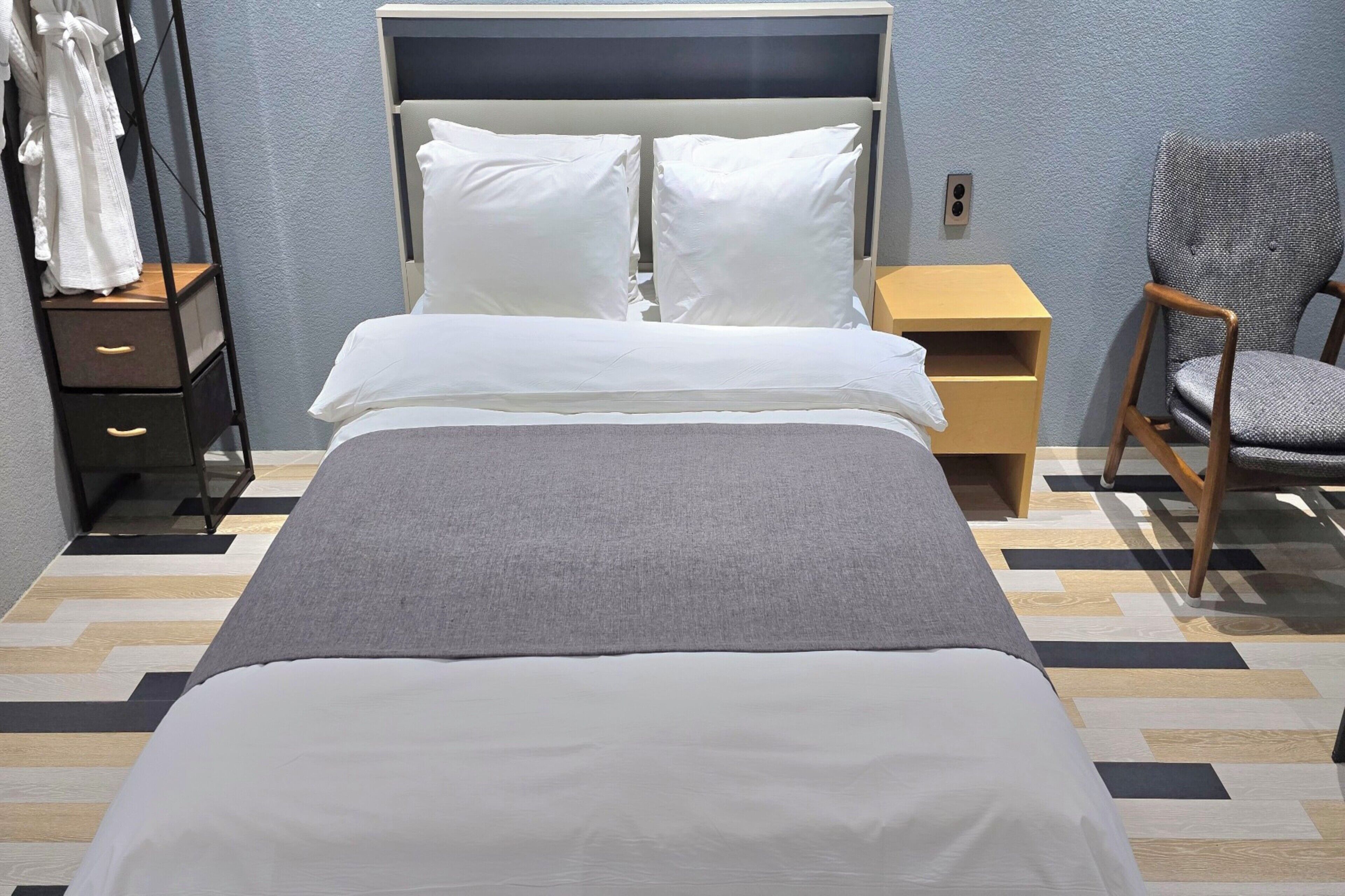 A neatly made bed with white bedding, a grey throw, and a wooden nightstand with a lamp.