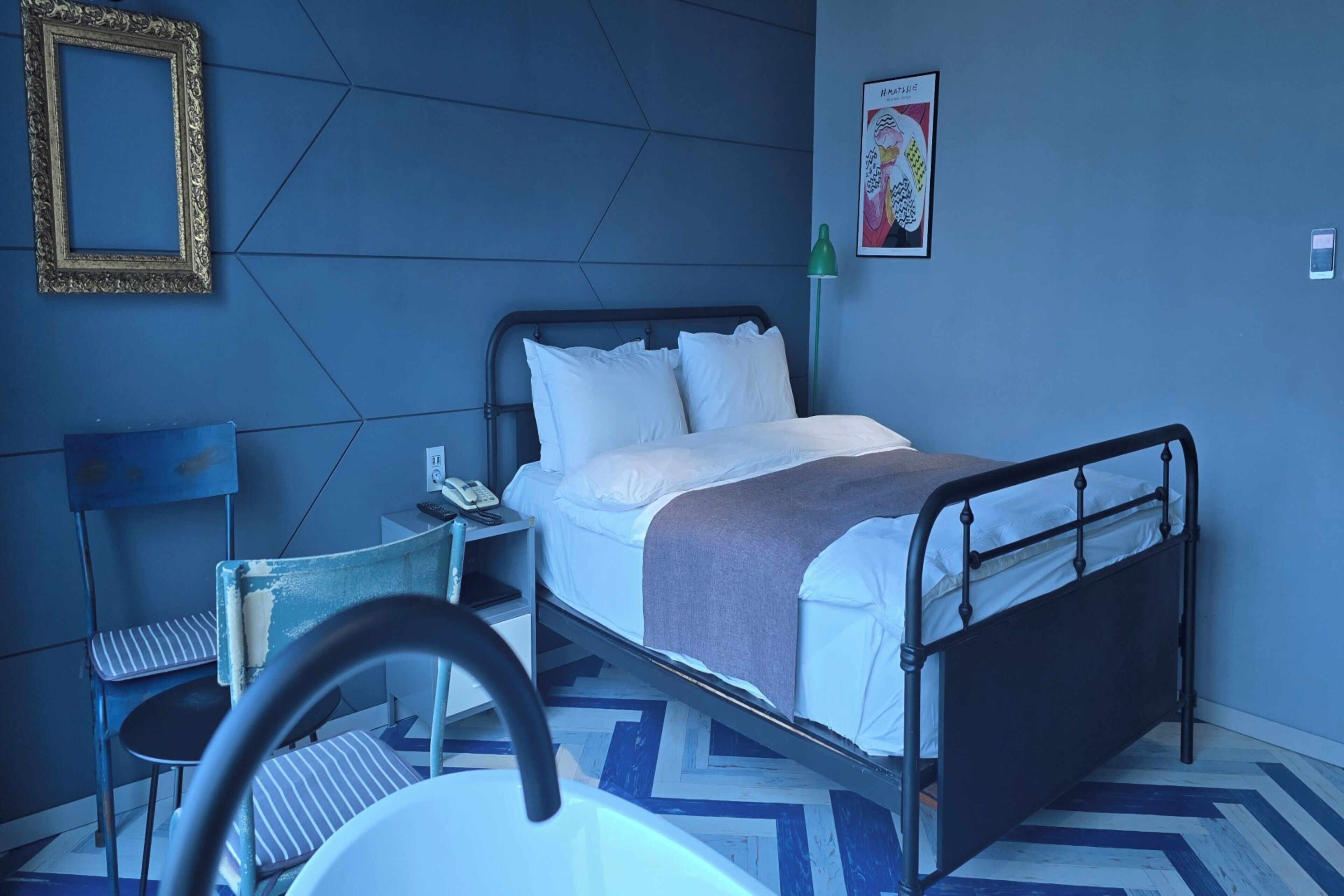 A room with a bed, a chair, a desk with a phone, and a picture on the wall.