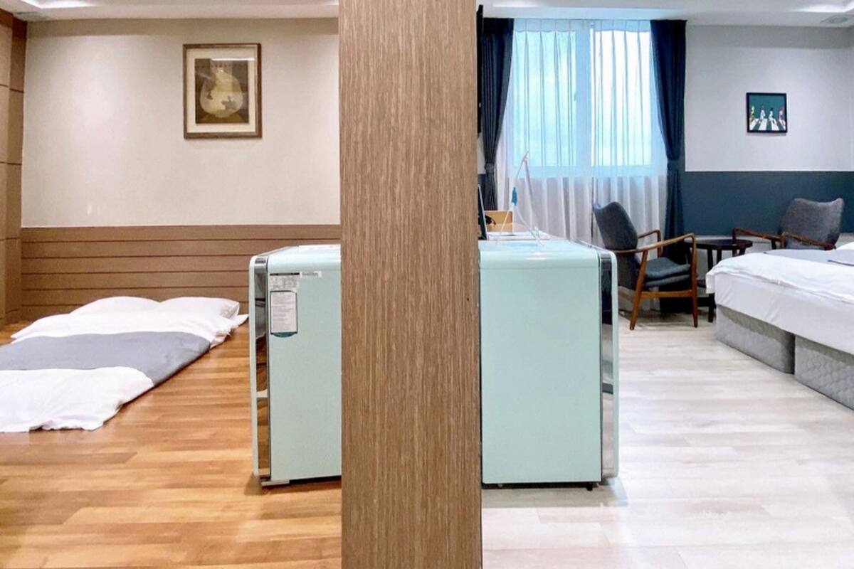 A hotel room with a bed, a desk, a chair, and a small table. There is a window with curtains and a painting on the wall.