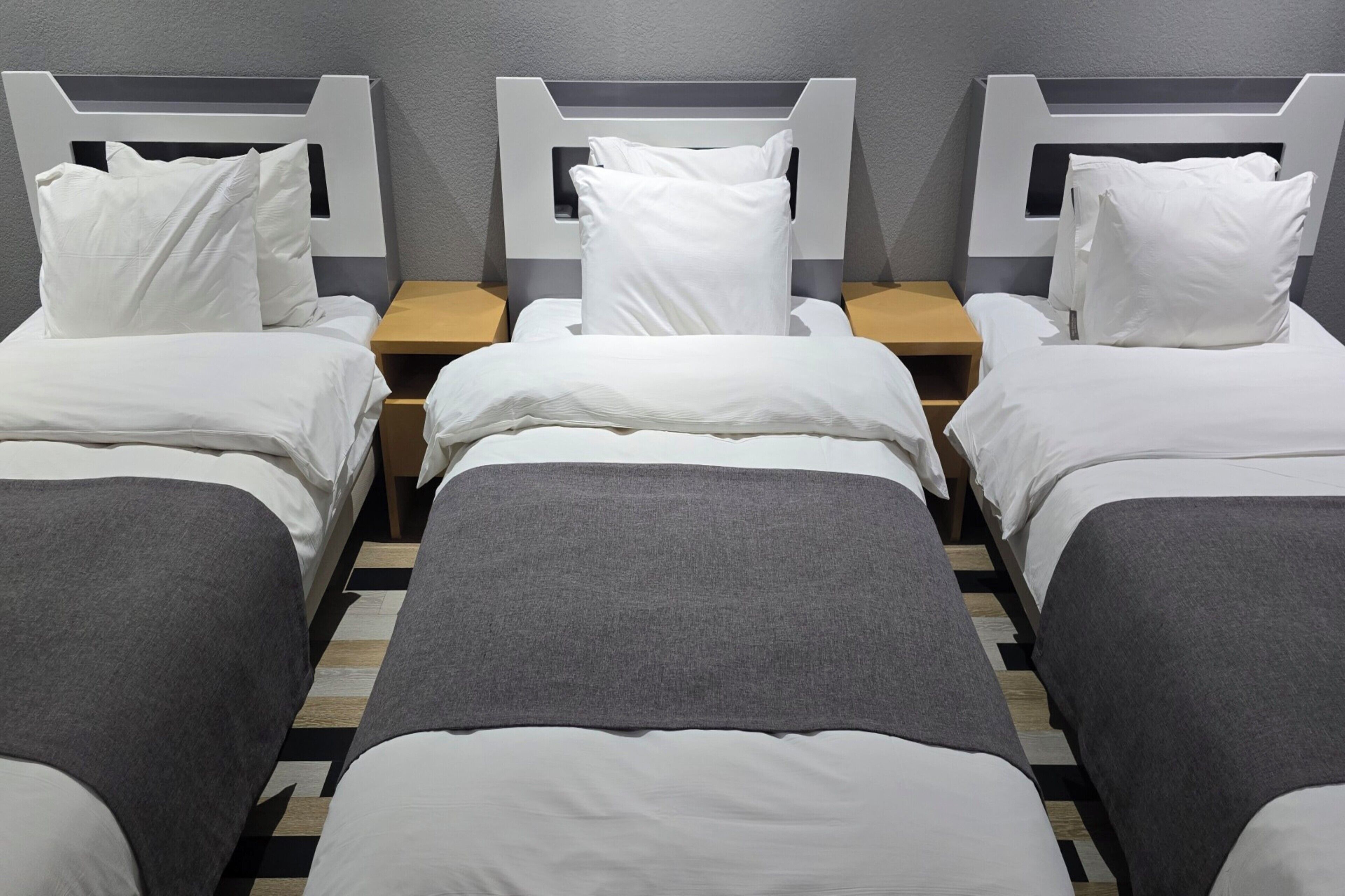 Une chambre d’hôtel avec trois lits simples, chacun doté de draps et d’oreillers blancs, ainsi que d’une couverture grise. Les lits sont disposés en rangée contre un mur gris, avec une table de chevet en bois placée entre chaque lit.