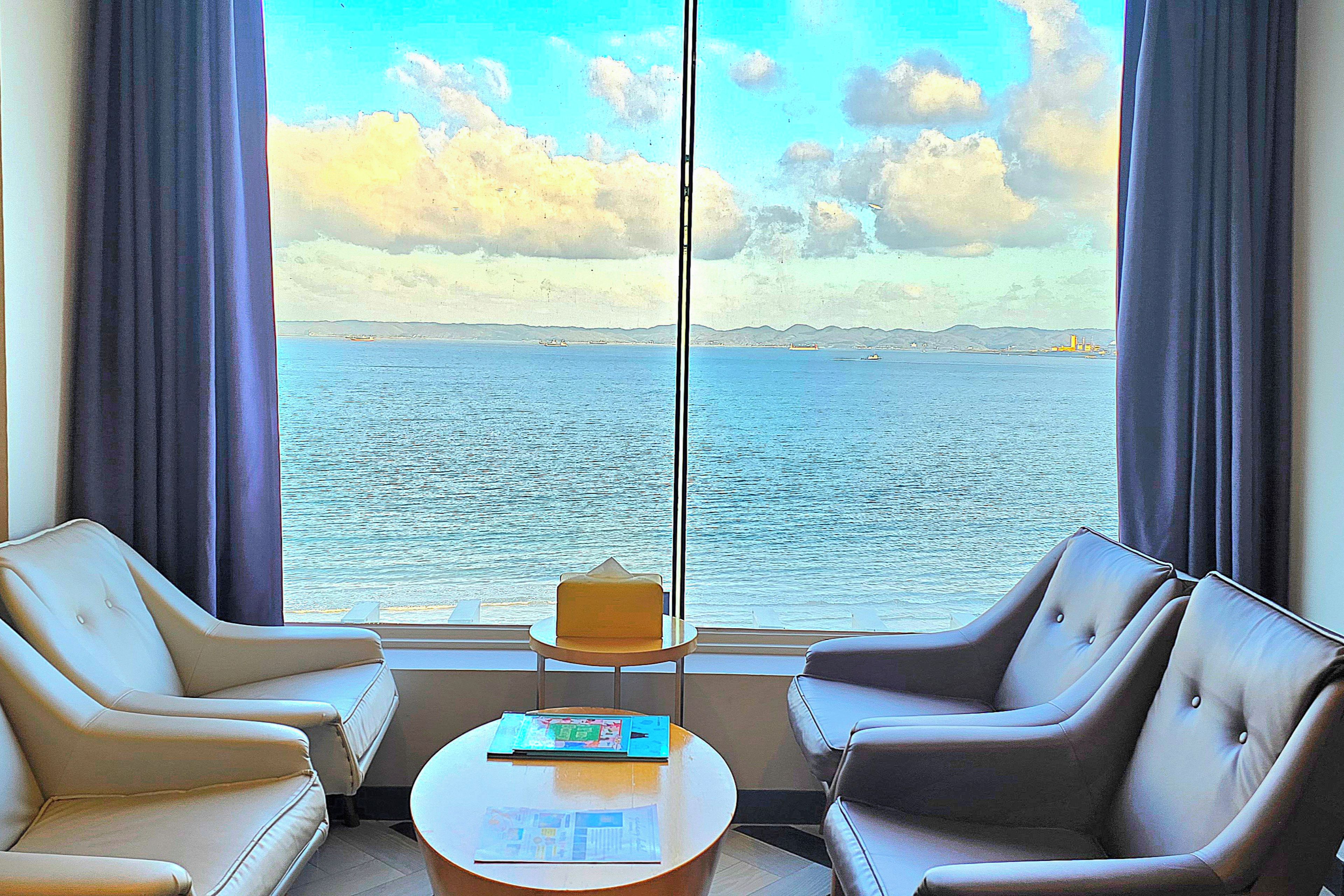 A room with two leather armchairs, a small table, and a view of the sea through large windows.