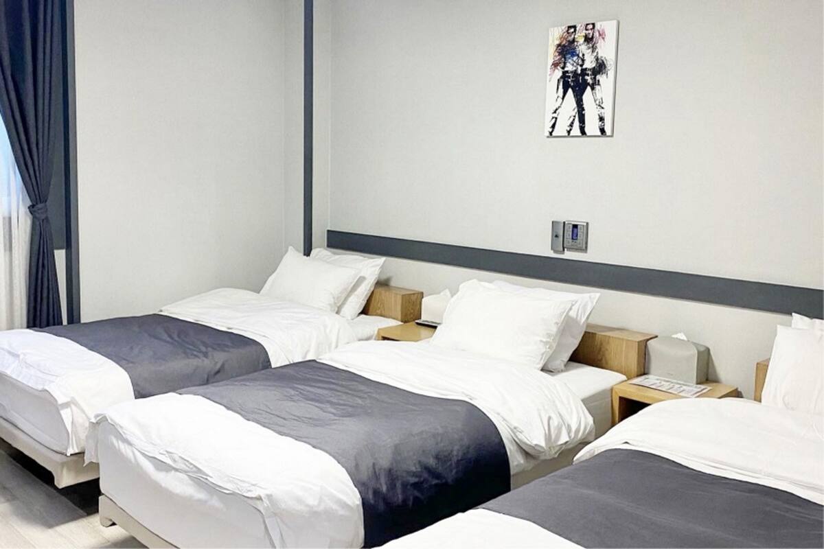 A hotel room with three single beds, each with white and grey bedding, wooden nightstands, and a wall-mounted light switch.