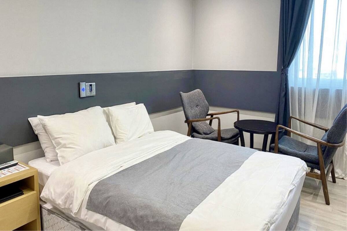 A hotel room with a large bed, two chairs, a small table, and a thermostat on the wall.