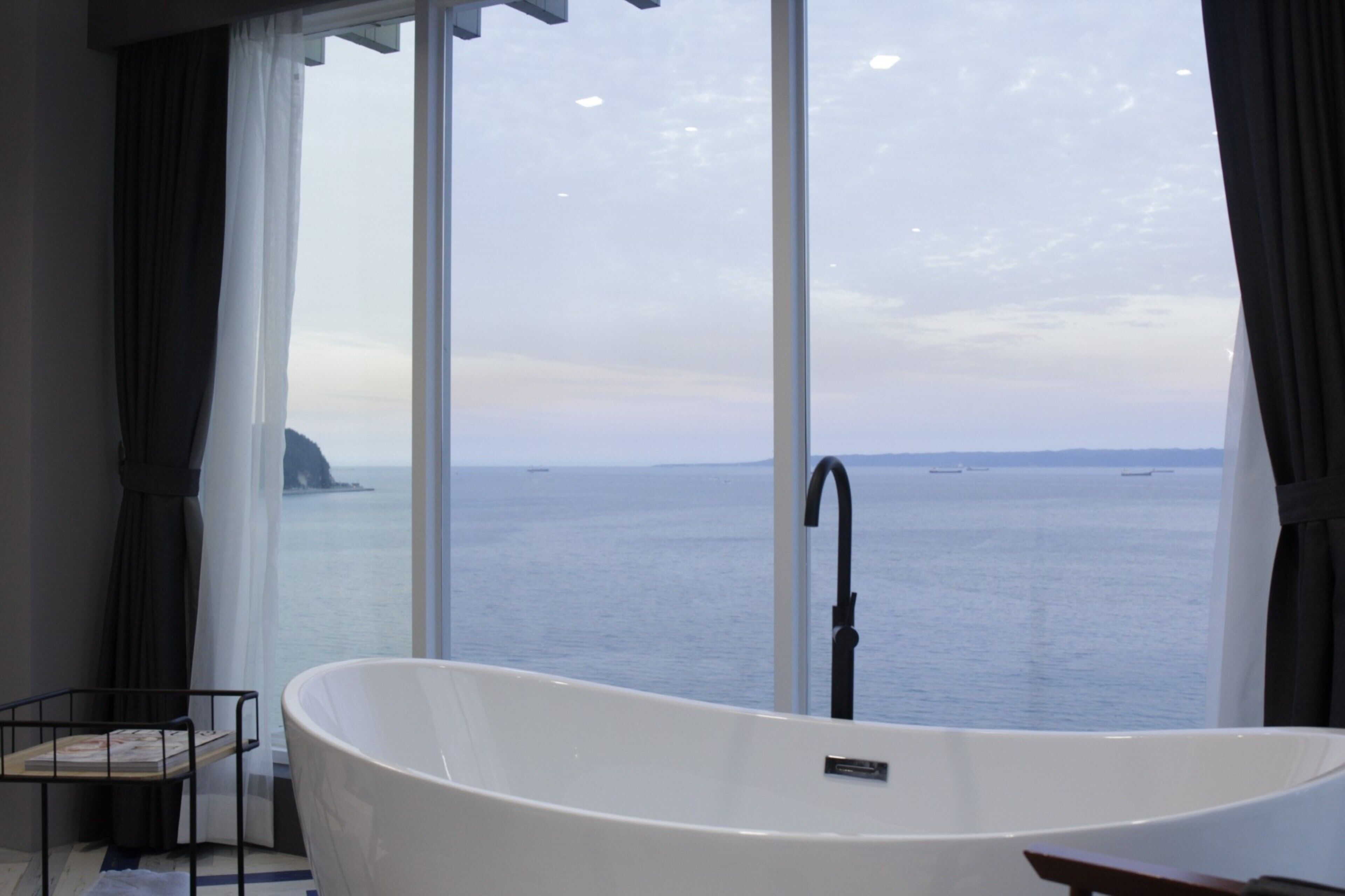A modern bathroom with a freestanding bathtub, a window with curtains, and a view of the sea.