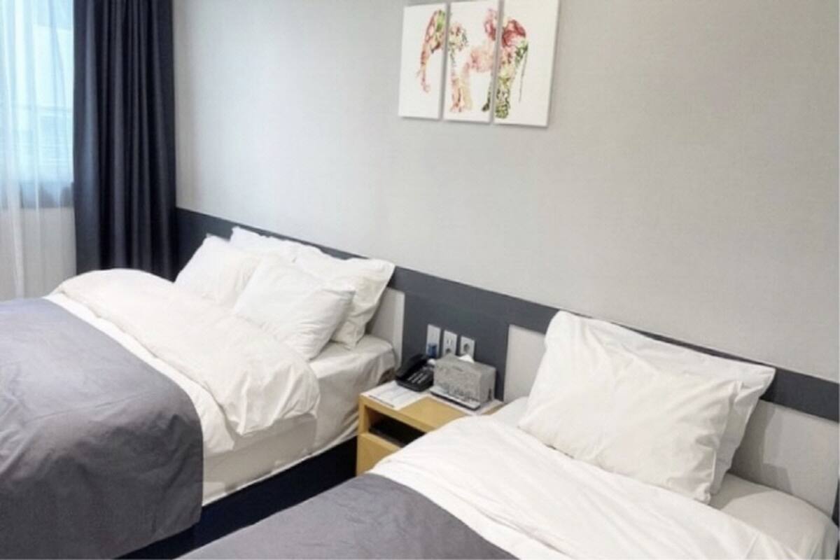 A hotel room with two beds, a bedside table with a phone and a lamp, and a wall with a floral artwork.