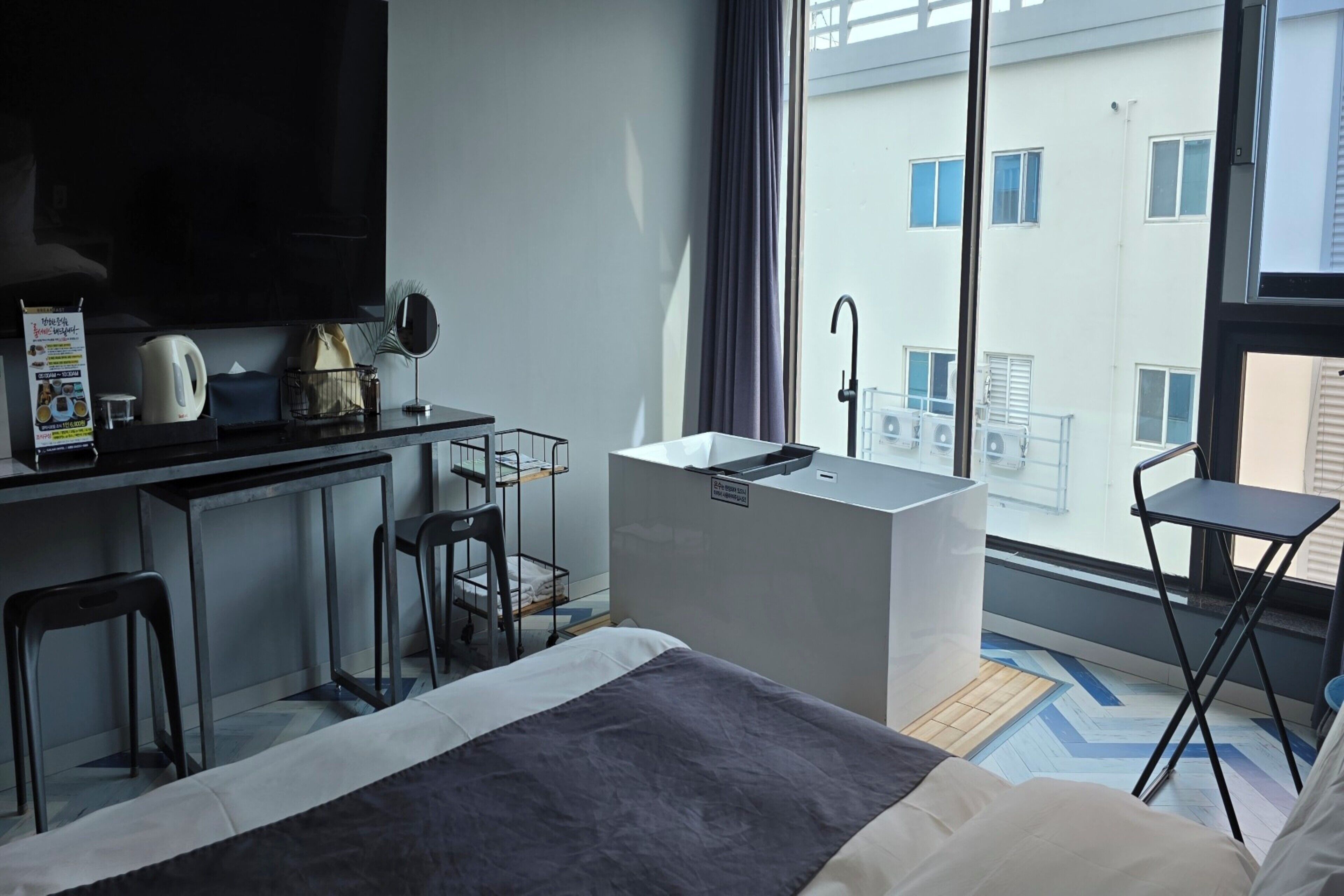 A modern hotel room with a bed, a desk with a kettle, a small kitchenette, and a view of the city through large windows.