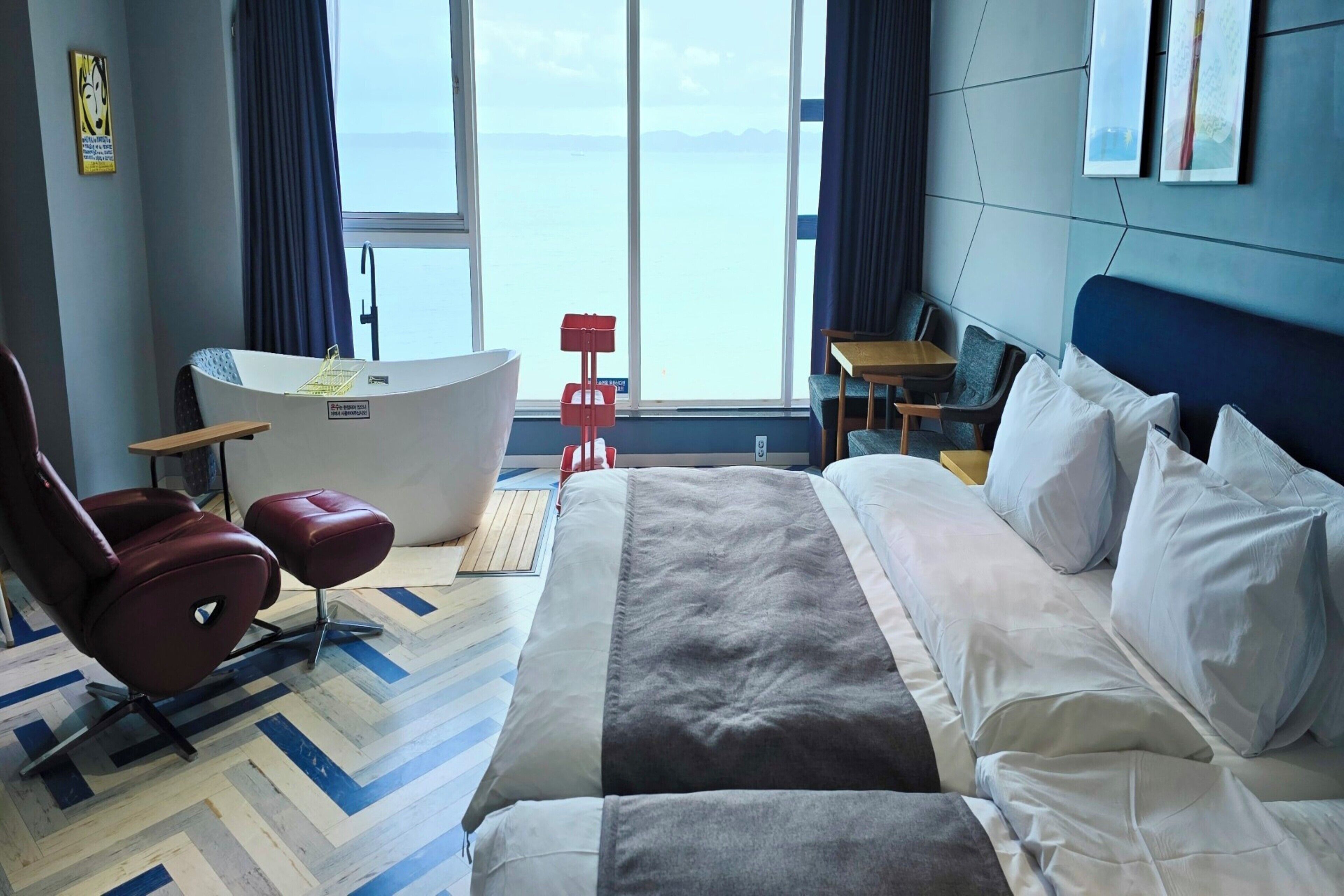 A hotel room with a bed, a desk, a chair, a bathtub, and a view of the sea.