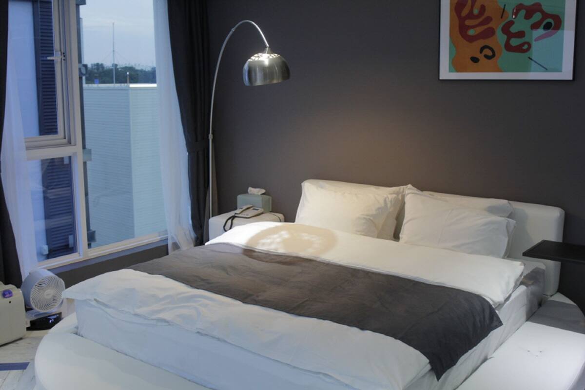 A hotel room with a bed, a bedside table, a lamp, and a window with blinds.