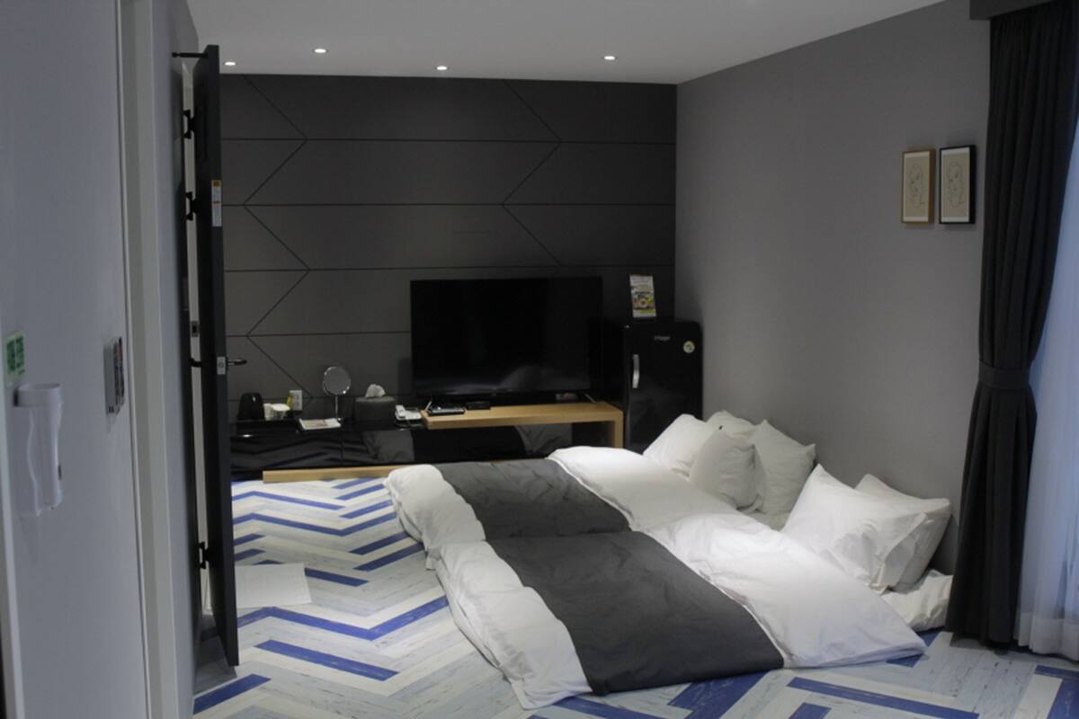 A modern hotel room with a large bed, a flat-screen TV, a desk with a computer, and a wall-mounted TV.