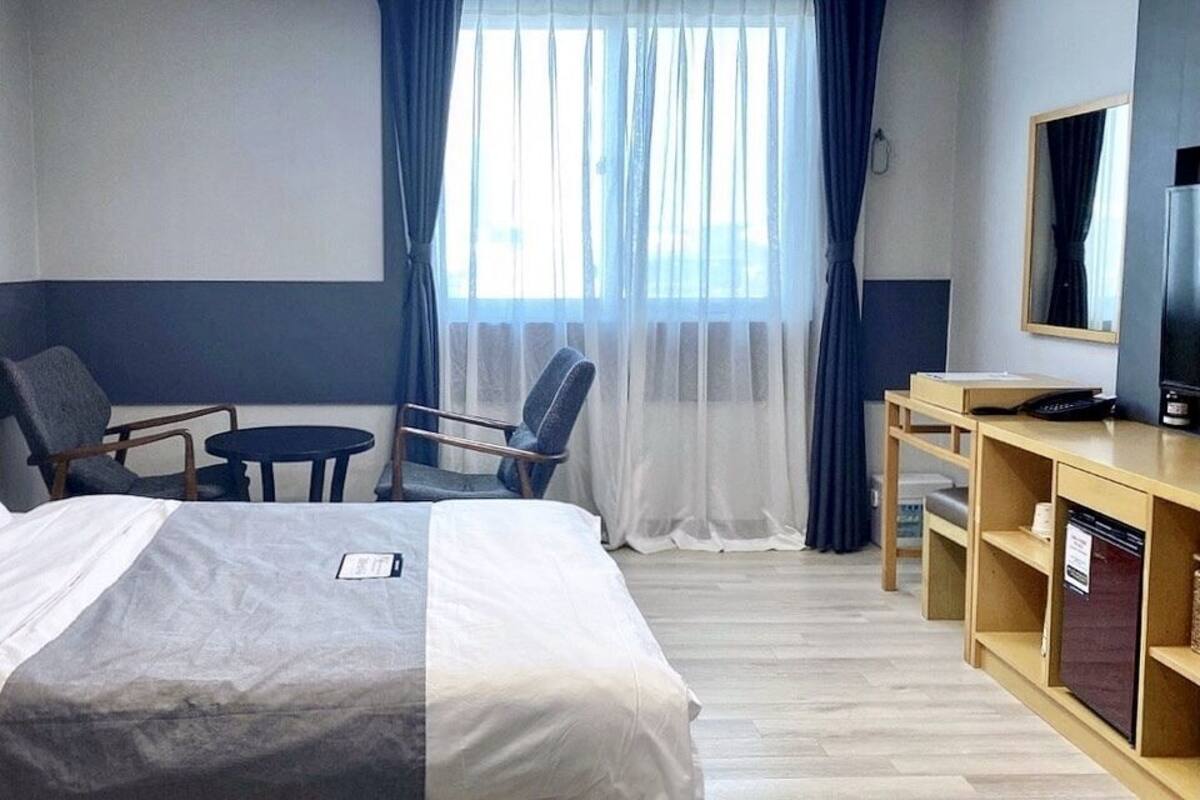 A hotel room with a bed, two chairs, a desk, a television, and a window with curtains.