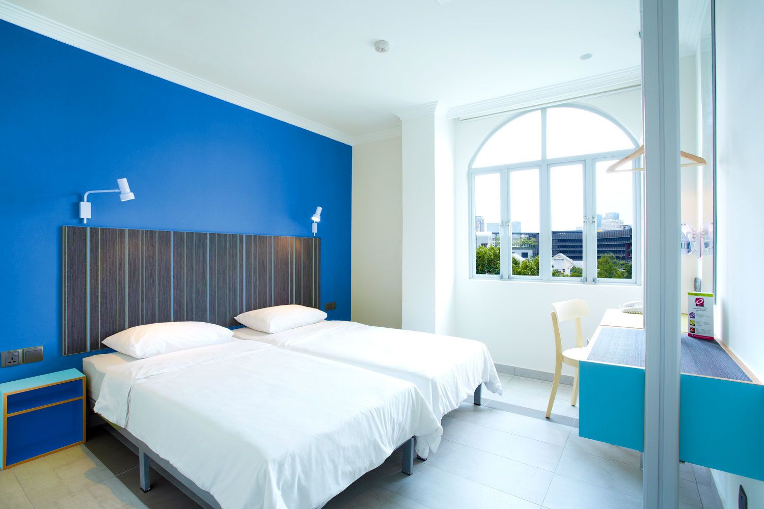 A modern hotel room with two beds, a desk, and a large window offering a city view.