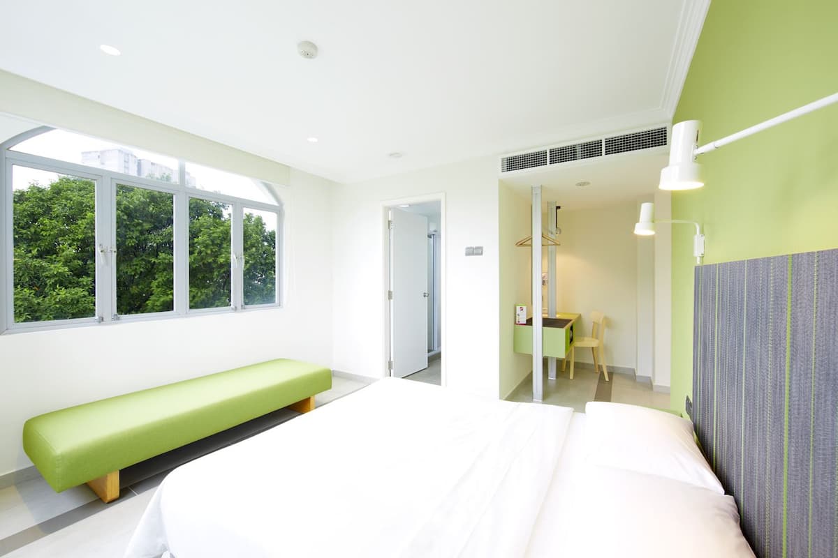 Une chambre d’hôtel moderne avec un grand lit, un banc vert, un bureau et une salle de bain visible à travers une porte ouverte.