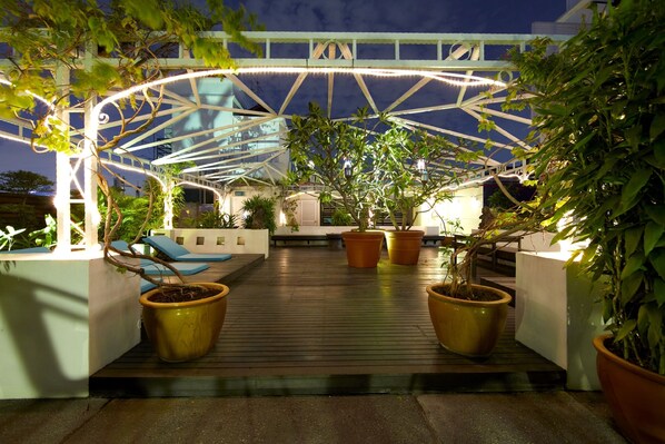 A covered outdoor area with wooden flooring, potted plants, and a white structure overhead.