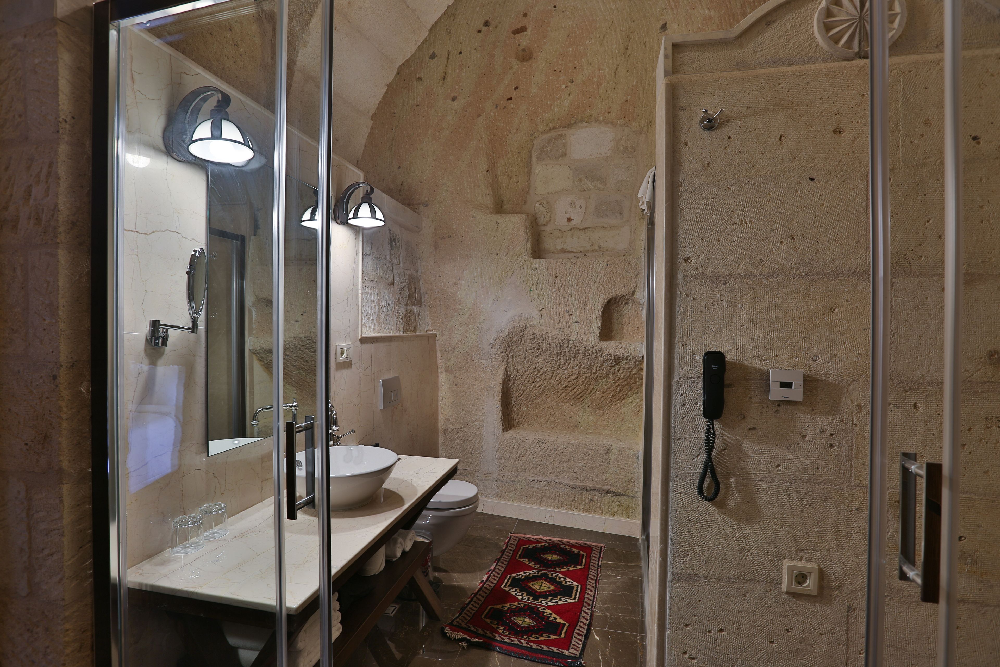 A bathroom with a glass shower enclosure, a sink with a mirror, a toilet, and a telephone.