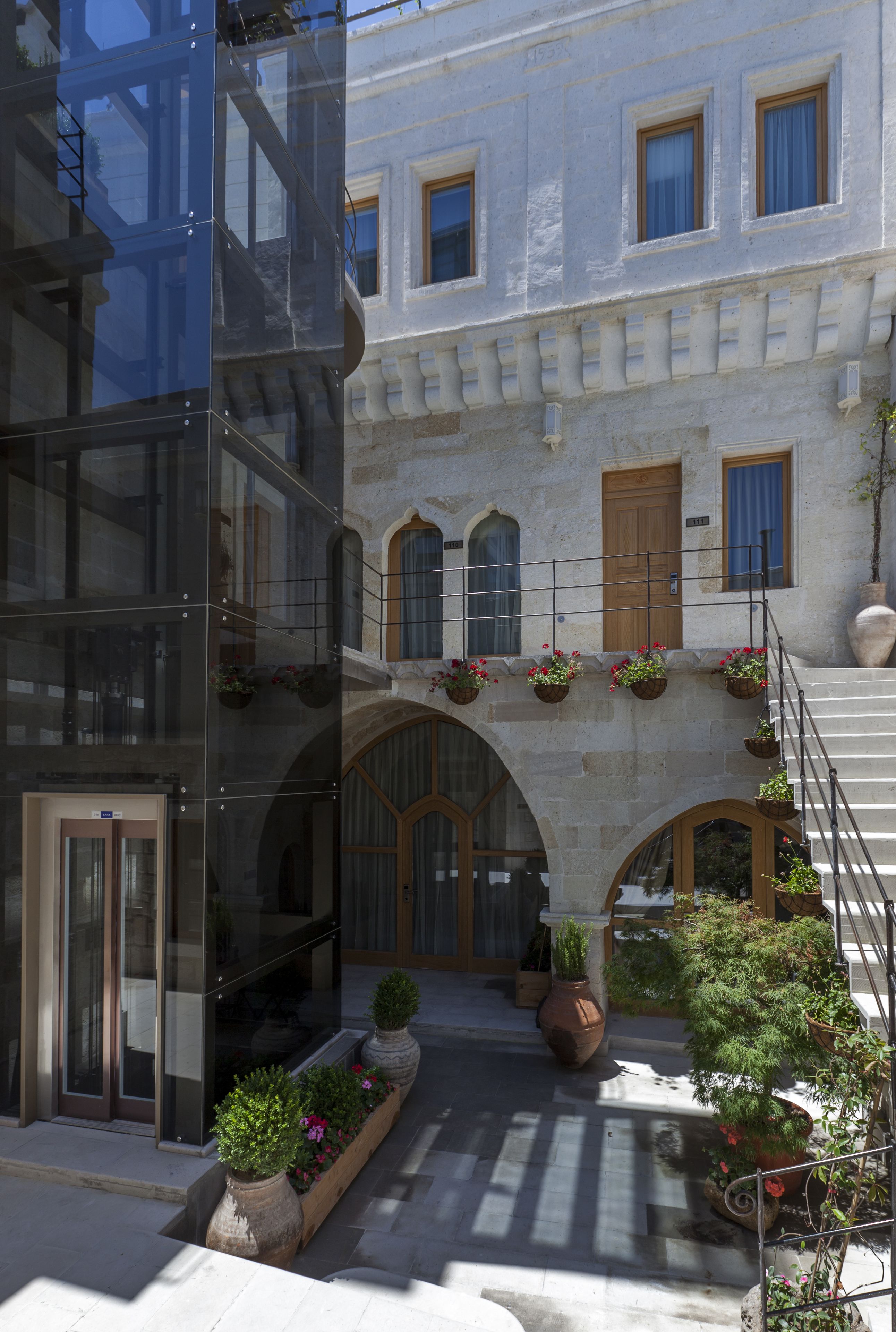 A courtyard with a modern glass building, stone arches, wooden doors, and potted plants.