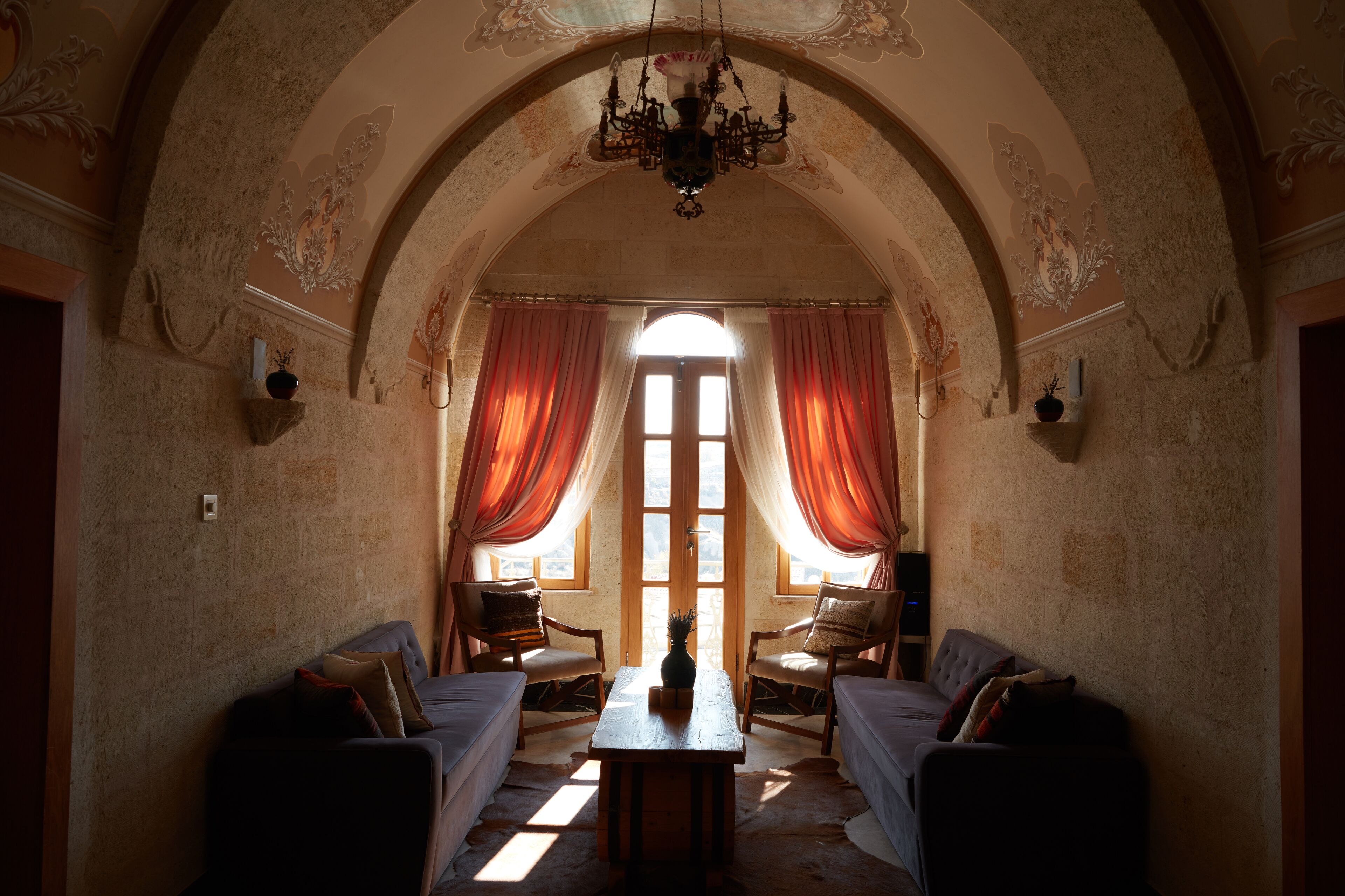 A room with a high arched ceiling, a chandelier, two armchairs, a sofa, a table, and a window with curtains.