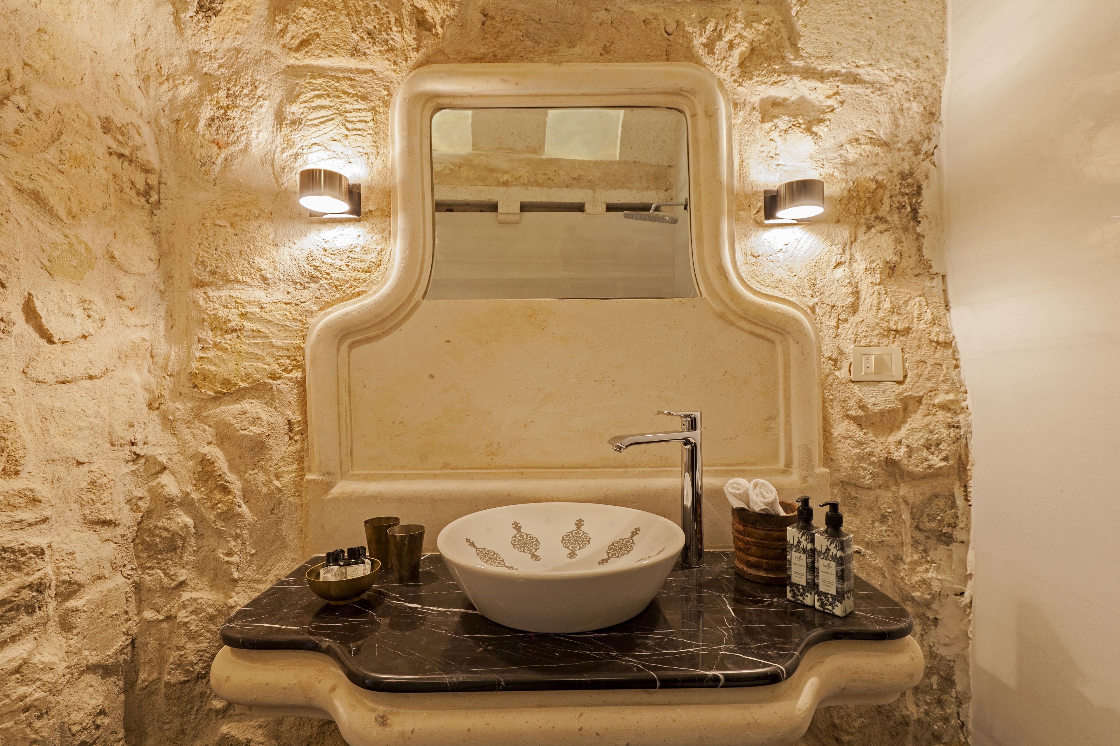 Un bagno con lavandino bianco, un piano di lavoro in marmo e una parete in pietra strutturata.