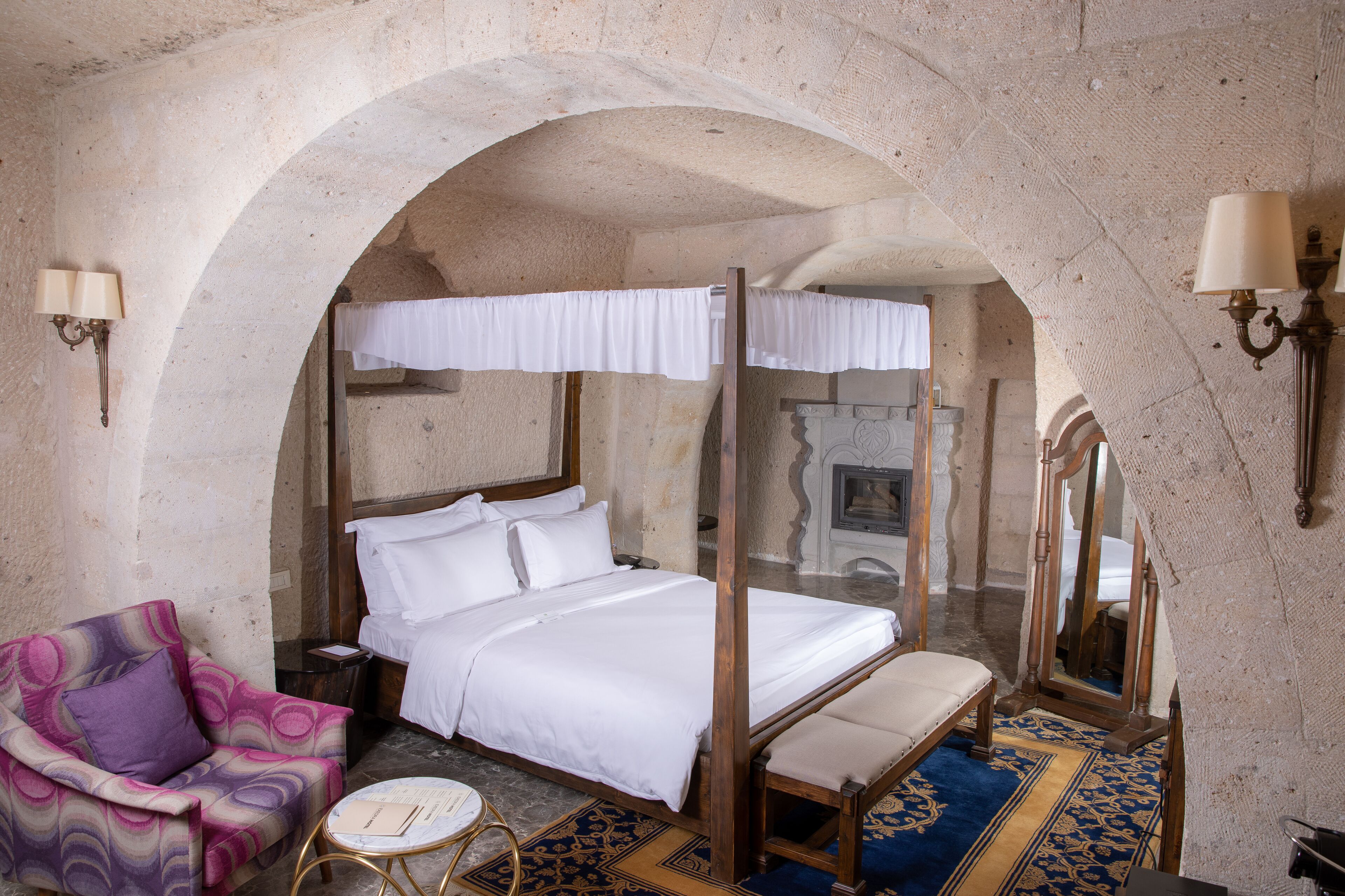 A four-poster bed with white linens, a wooden bench, a purple armchair, and a small round table with a book.