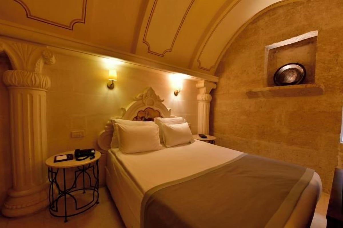 A hotel room with a bed, a small round table, and a shelf with a bowl.