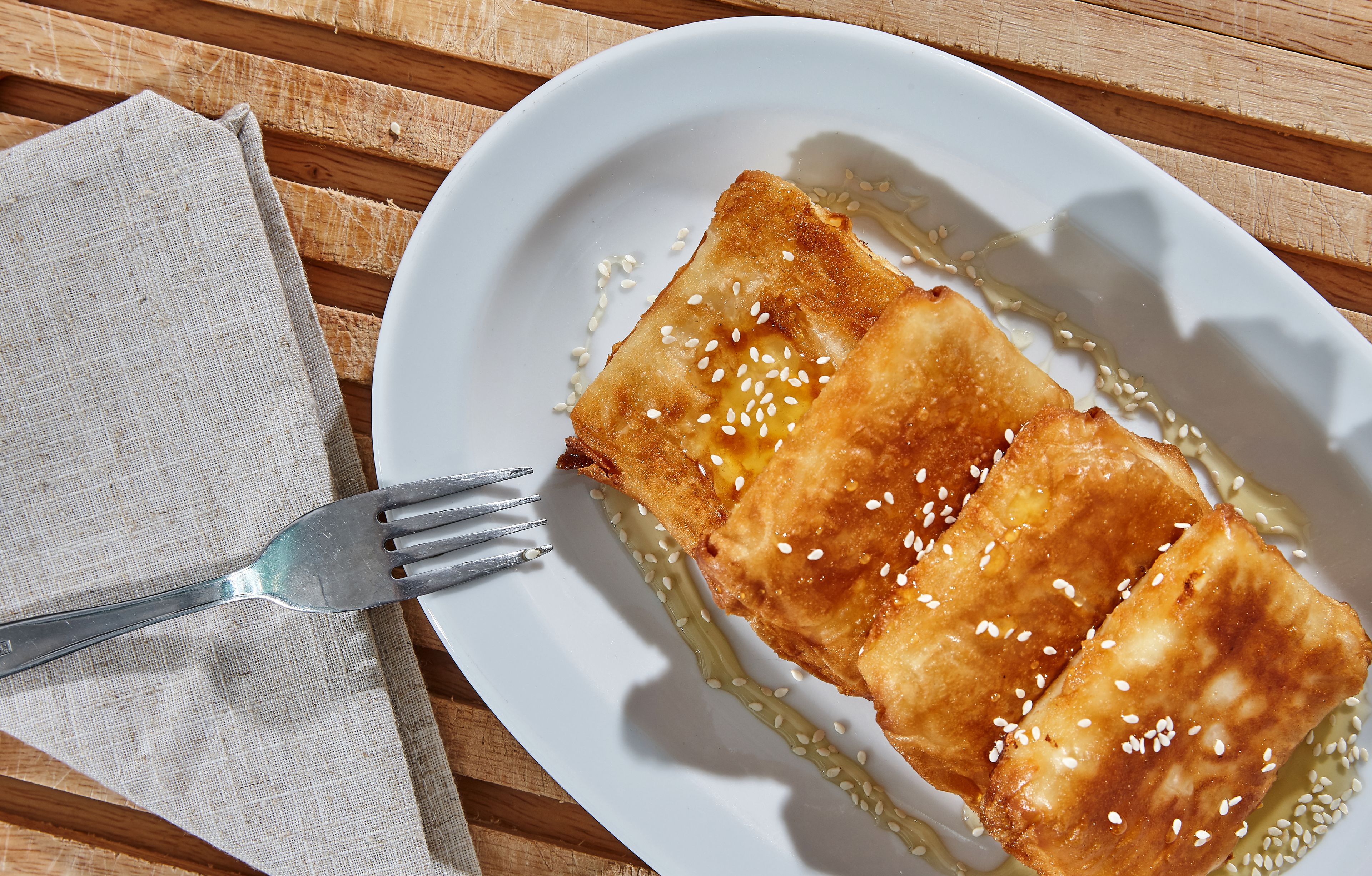Un plato con dos porciones de comida frita adornada con semillas de sésamo, acompañada de un tenedor y una servilleta sobre una mesa de madera.