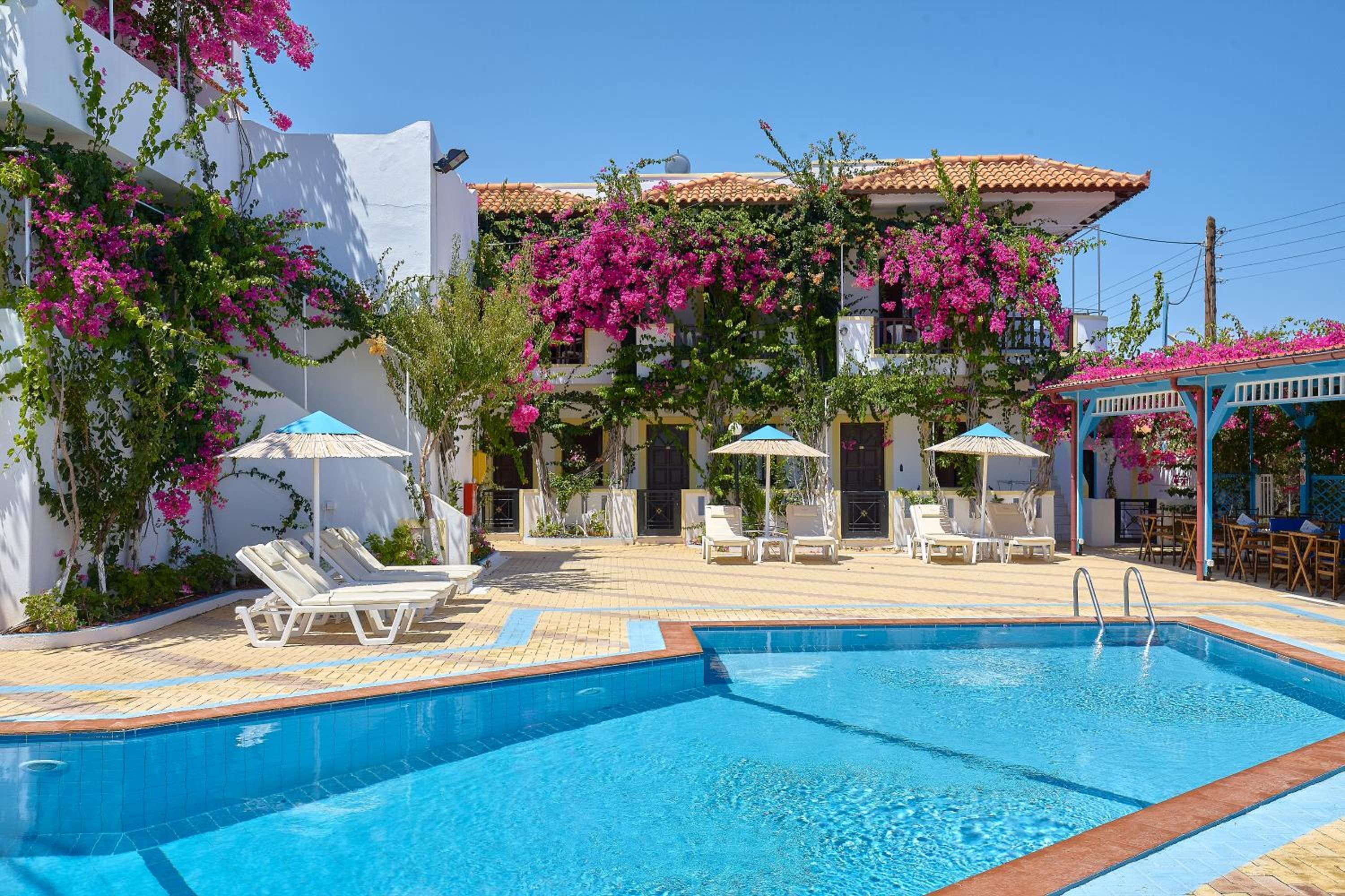 A swimming pool with lounge chairs and umbrellas, surrounded by a building with pink flowering vines.