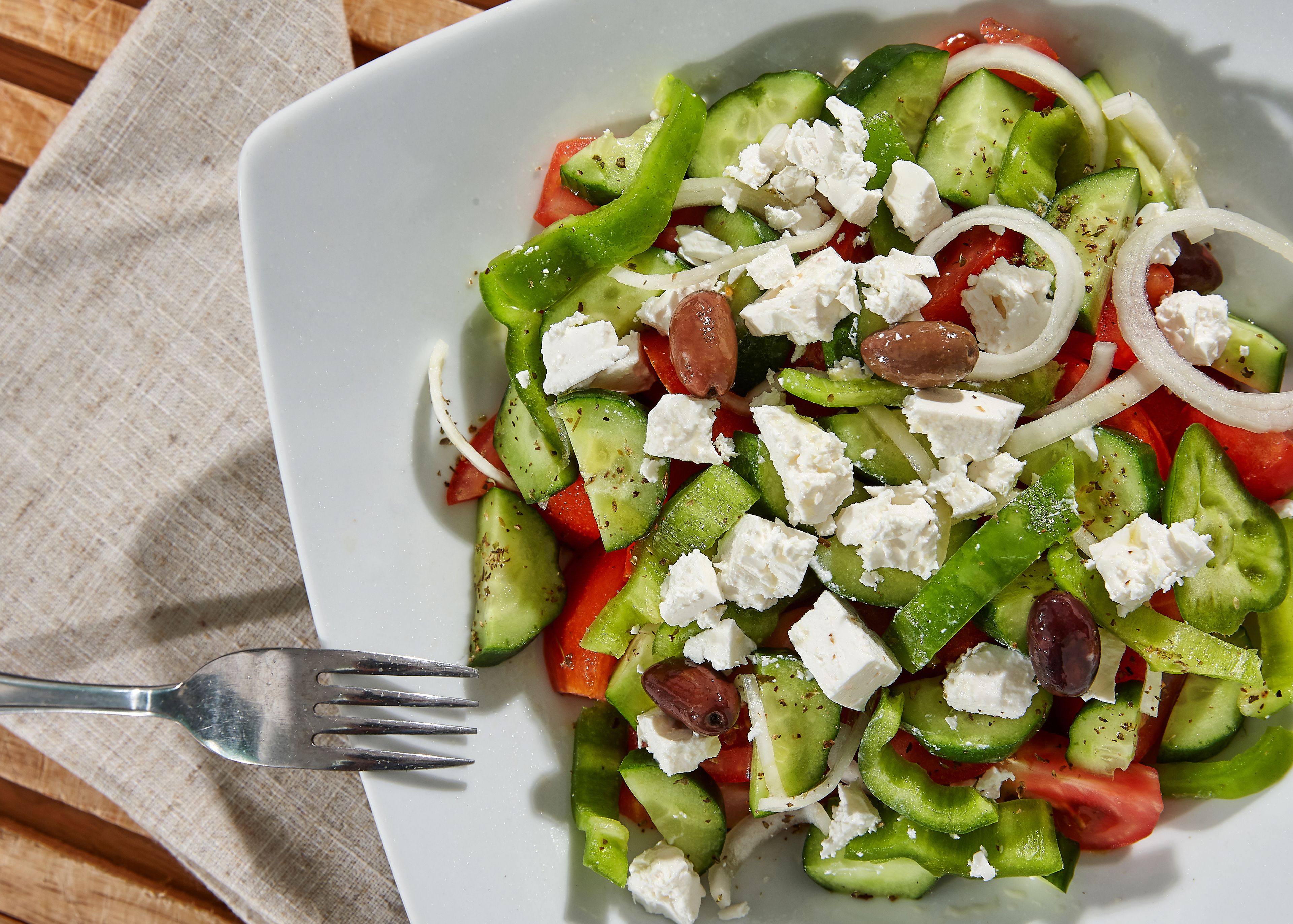 A white plate with a Greek salad containing cucumbers, tomatoes, olives, and feta cheese, accompanied by a fork on a wooden table.
