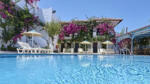 Piscina com espreguiçadeiras e guarda-sóis, cercada por edifícios com varandas e plantas floridas.