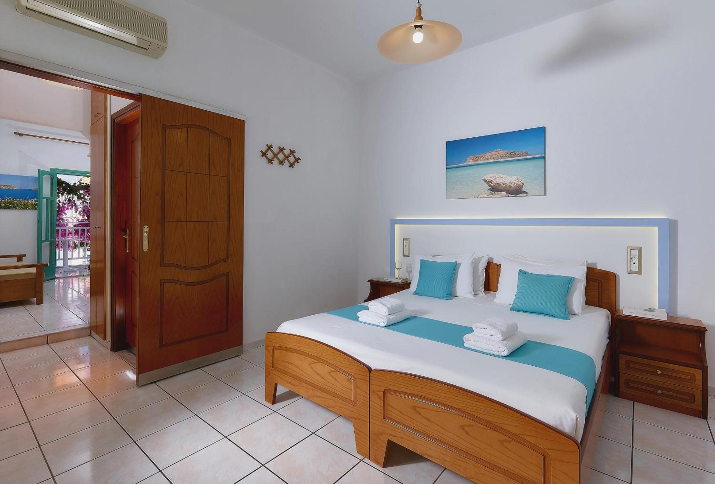 A hotel room with a wooden bed, a nightstand, a wall-mounted lamp, a painting of a beach, and a large wooden door.