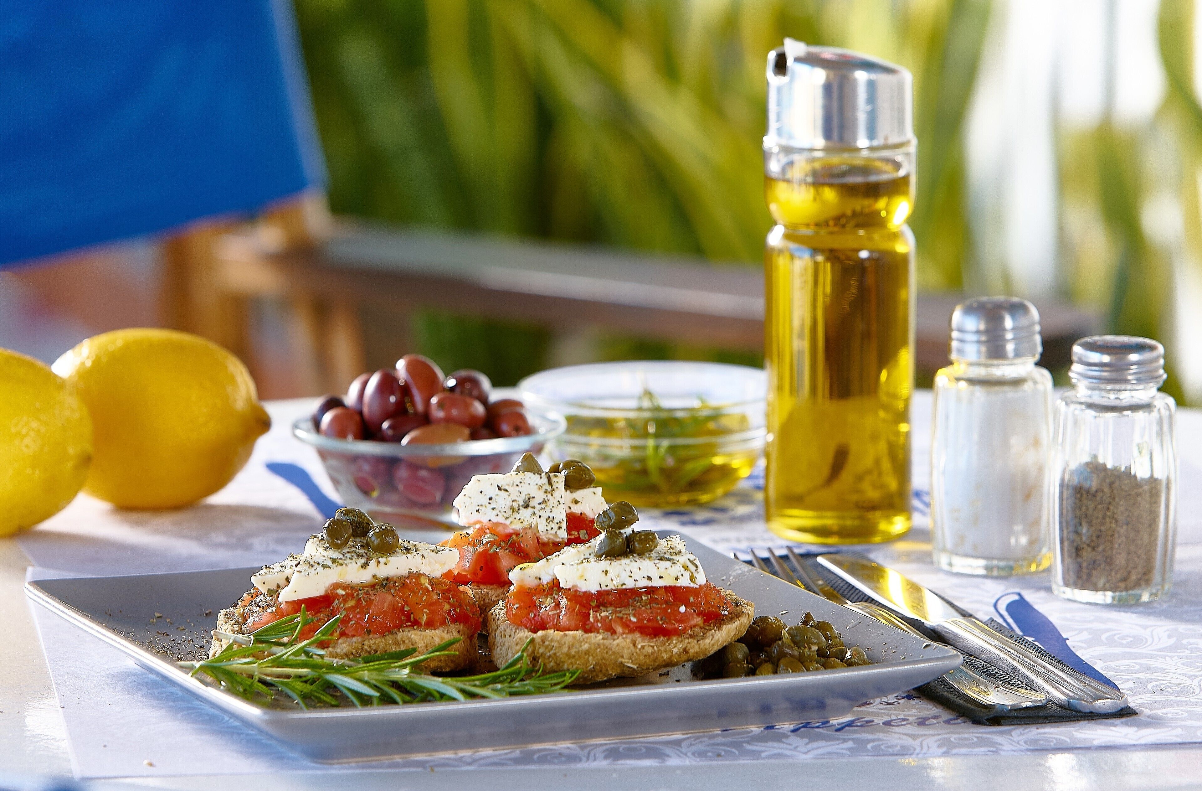 Un plato de bruschetta con tomates, aceitunas y hierbas, acompañado de saleros y pimenteros, y aceite de oliva sobre una mesa.