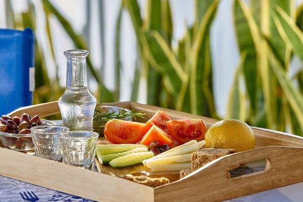 A wooden tray with a variety of fruits, cheese, and olives, accompanied by a glass of water and a bottle, set against a backdrop of green plants.