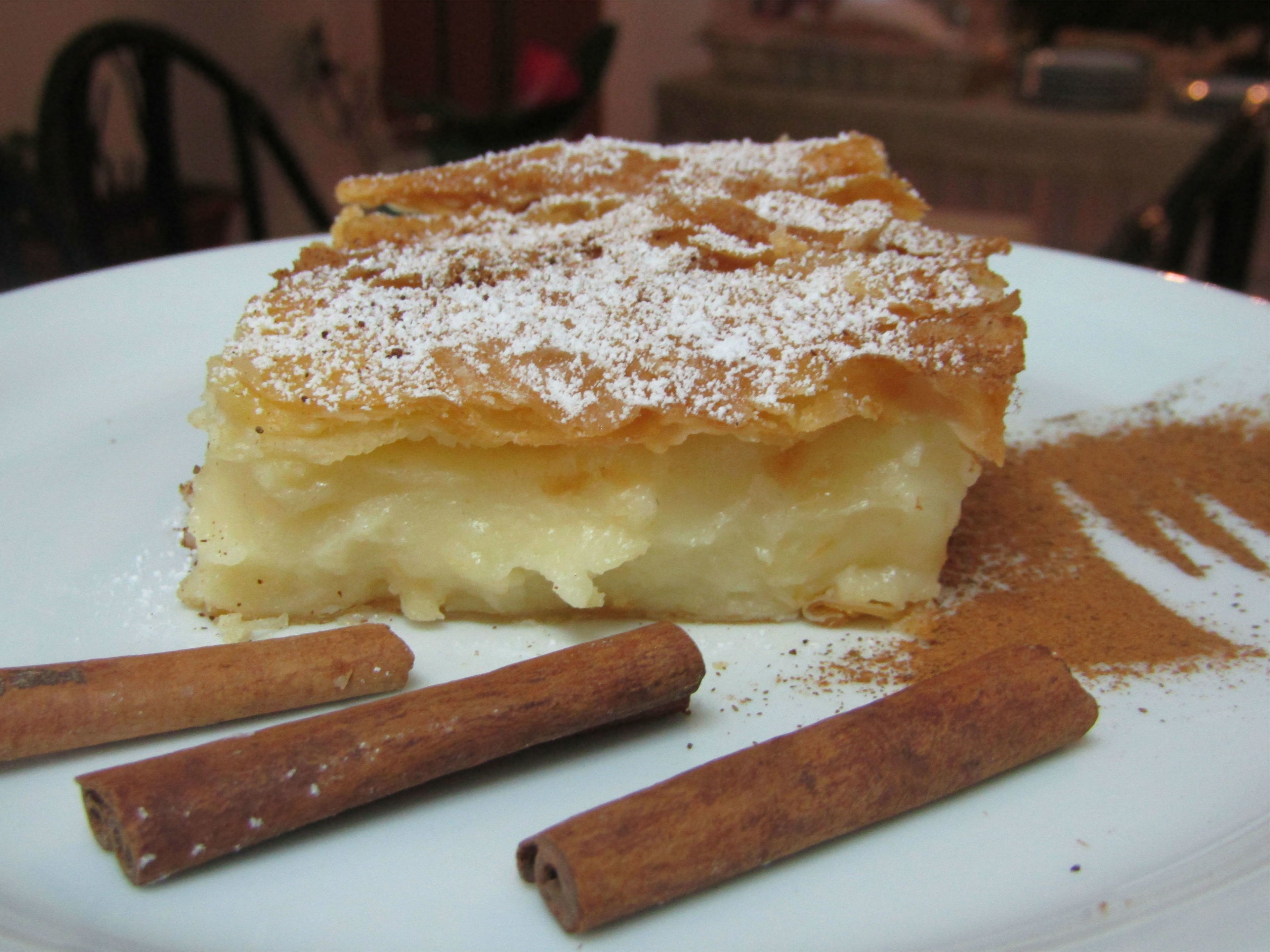 Una porción de tarta de manzana en un plato blanco con palitos de canela al lado.