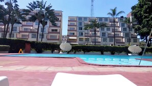 A swimming pool with lounge chairs, palm trees, and a multi-story building in the background.