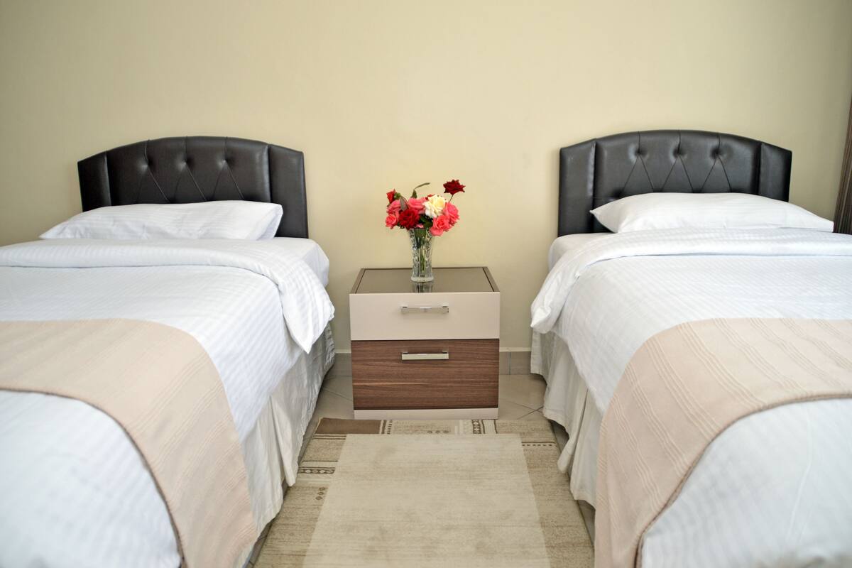 A hotel room with two beds, a nightstand with a vase of flowers, and a beige carpet.
