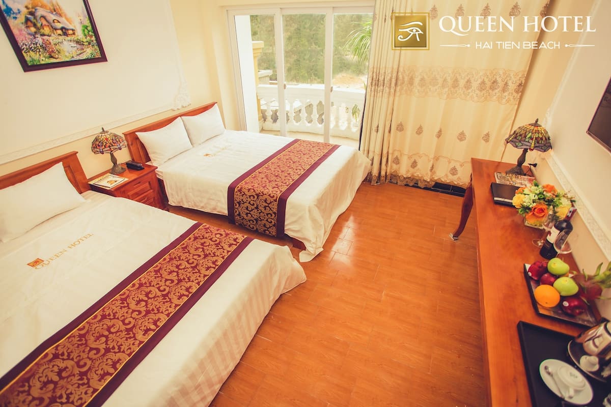 Habitación de hotel con dos camas, suelo de madera, balcón con vistas y un escritorio con un jarrón de flores.