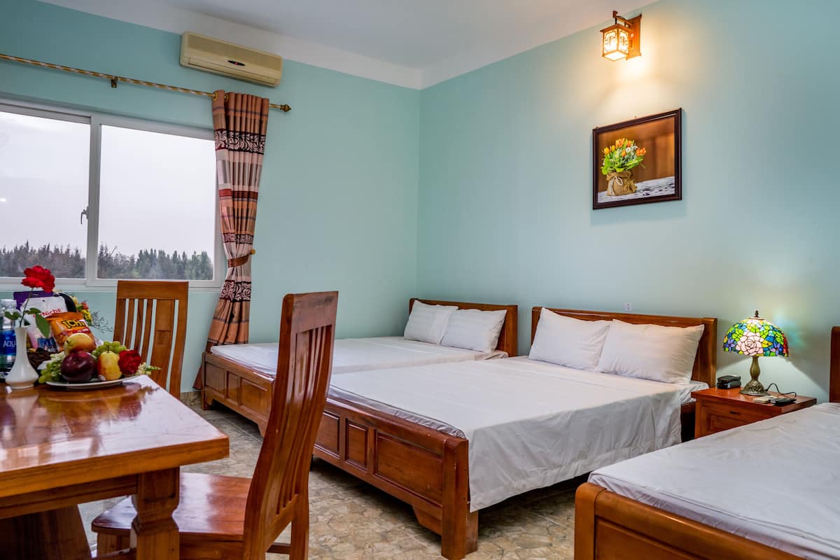 Una habitación con dos camas, una mesa de comedor con fruta, una silla, una ventana con cortinas, un aire acondicionado de pared y una lámpara de pared.