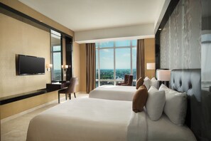 Habitación de hotel con dos camas, un escritorio, un televisor y un ventanal con vistas a la ciudad.
