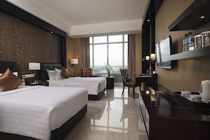 Habitación de hotel con dos camas, un escritorio, un televisor y un amplio ventanal con vistas a la ciudad.