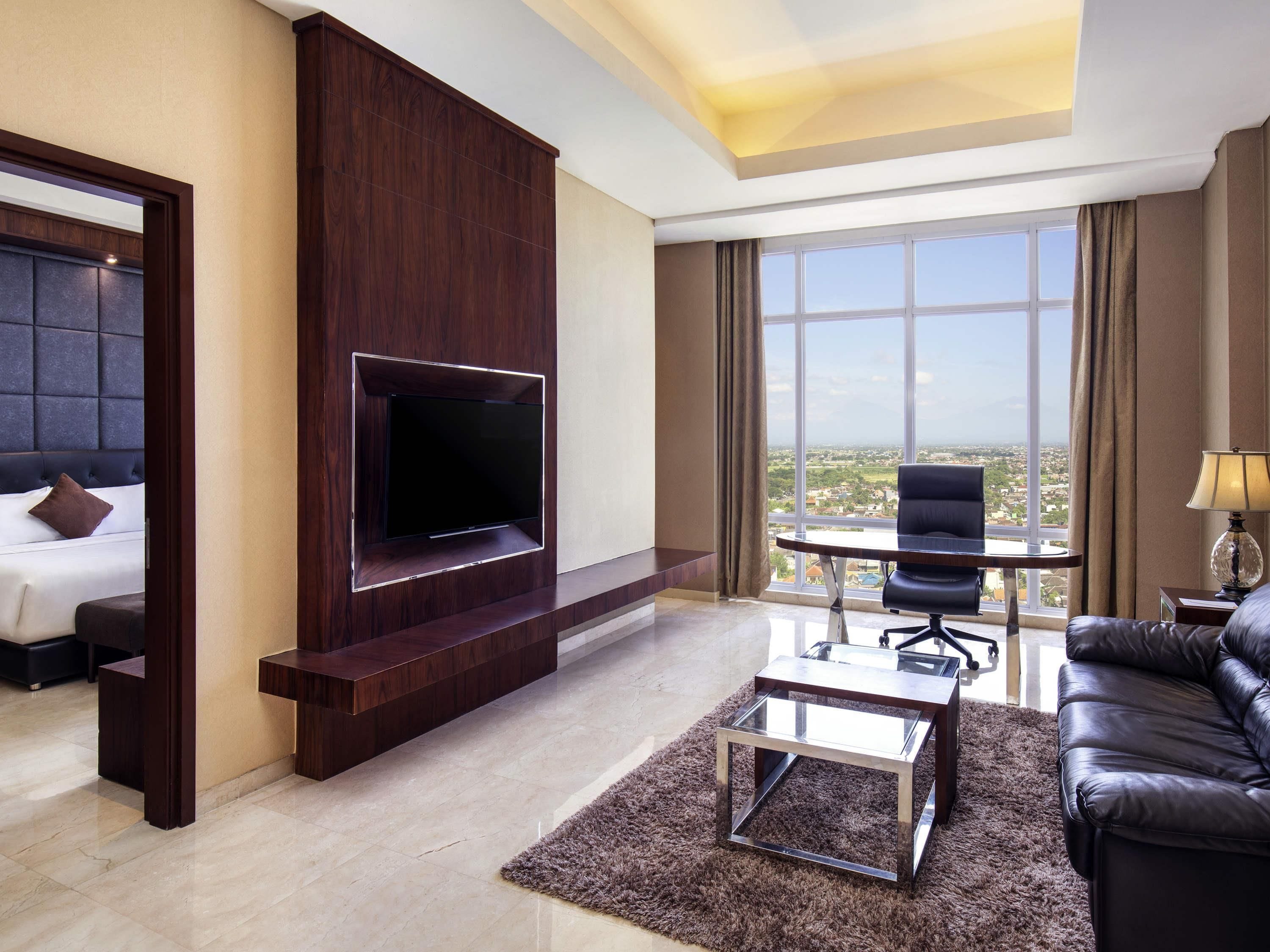 Habitación de hotel con televisión instalada en un panel de madera, cama con cabecera, sofá, una mesita y vista a la ciudad a través de amplias ventanas.