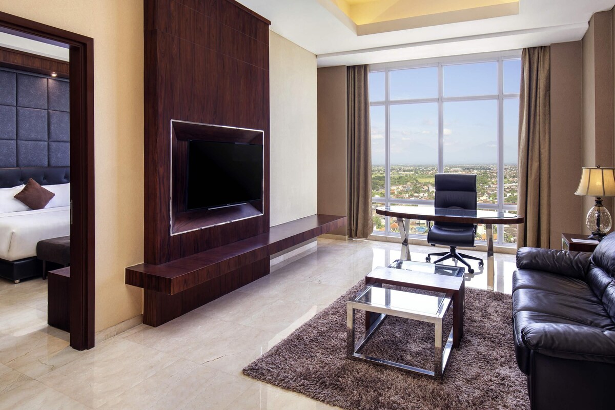 A hotel room with a TV mounted on a wooden panel, a bed with a headboard, a sofa, a small table, and a view of the city through large windows.