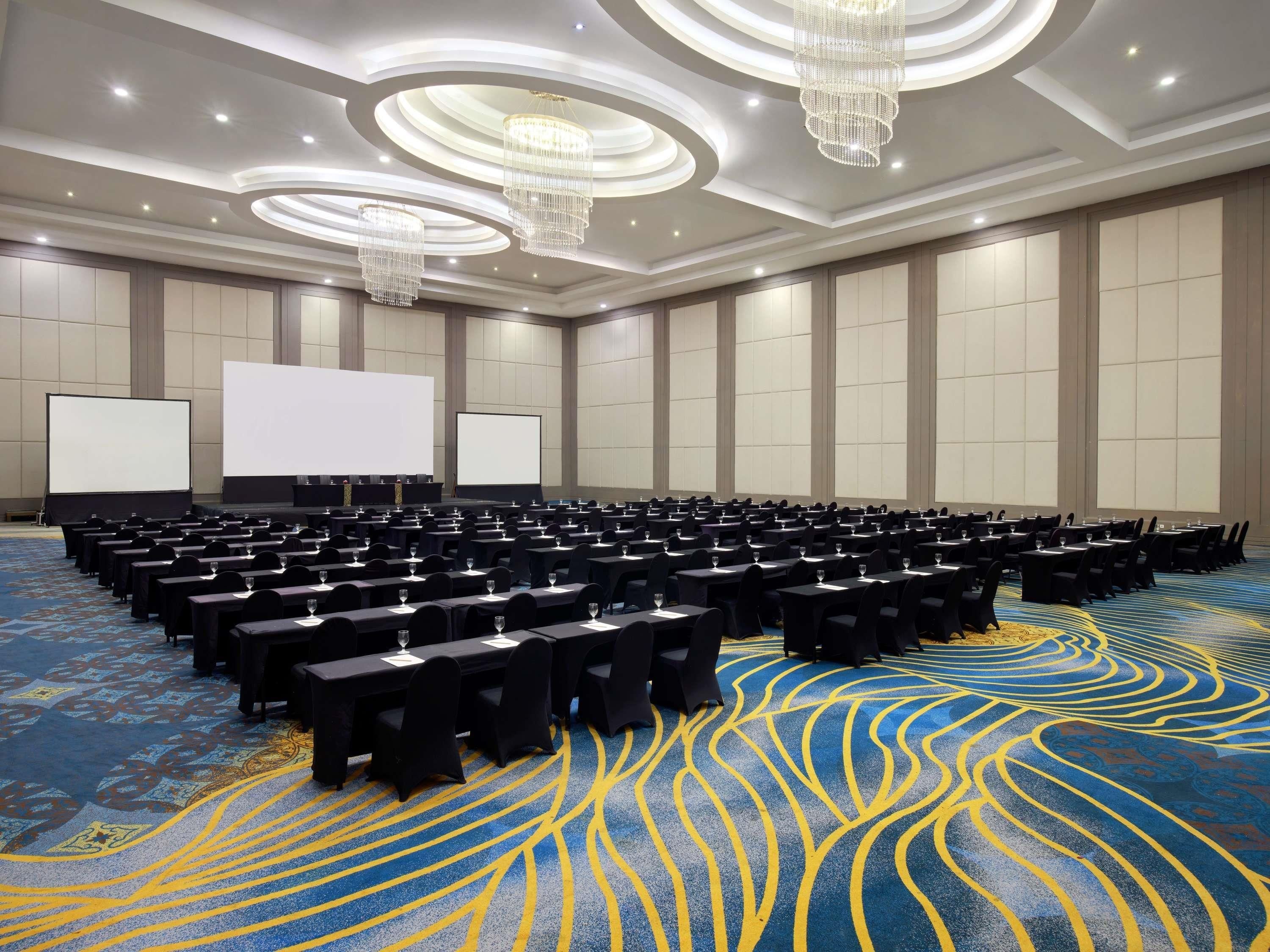 Un salón de conferencias con filas de mesas y sillas, una pantalla grande y una alfombra estampada.