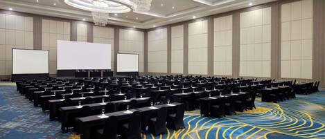 Un salón de conferencias con filas de mesas y sillas, una pantalla grande y una alfombra estampada.